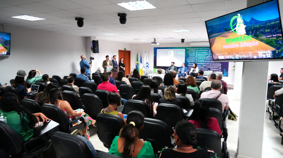A imagem mostra uma ampla sala com várias fileiras de cadeiras pretas ocupadas por participantes do Seminário Mosaico Jalação. No palco central, um grupo de palestrantes conduz a programação, enquanto o público, atento, acompanha as falas e apresentações.