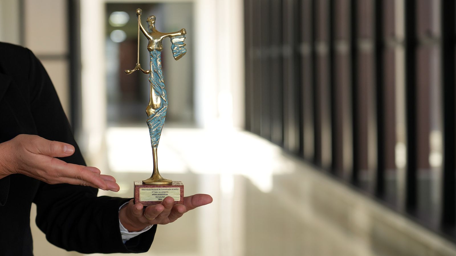 A imagem mostra uma pessoa vestindo um blazer preto segurando um troféu estilizado da deusa da Justiça (Têmis). A estatueta é dourada com detalhes em azul, representando a figura feminina em pé, segurando uma balança na mão esquerda erguida. A base do troféu é retangular, com uma pequena placa metálica contendo inscrições. O fundo está desfocado, revelando um corredor iluminado com janelas enfileiradas à direita e paredes claras, transmitindo um ambiente institucional.