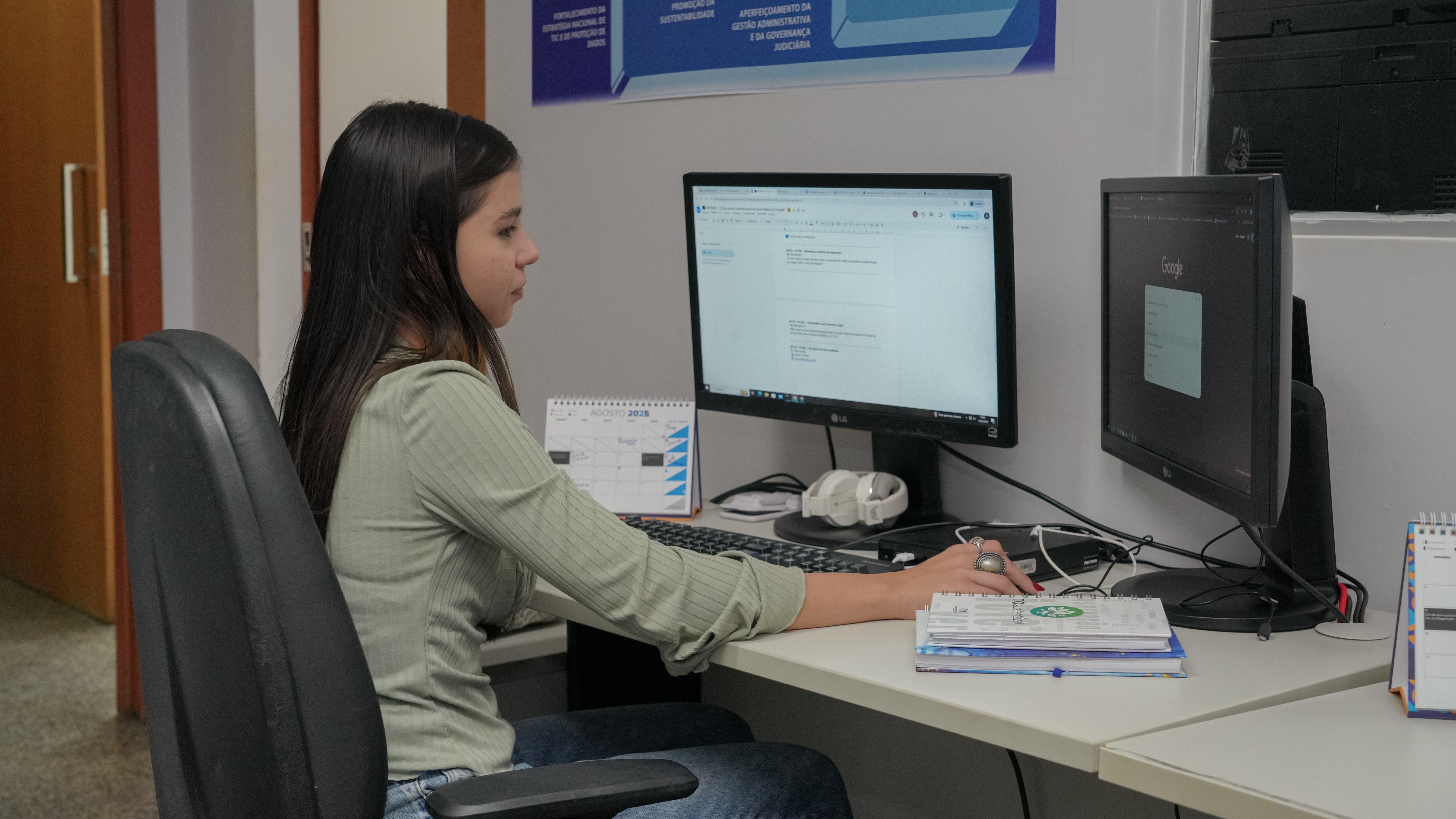 Foto mostra uma jovem sentada em frente a um computador, em um ambiente de escritório. A jovem está voltada para o computador, interagindo com ele. Ela está sentada em uma cadeira de escritório. O cenário é composto por uma mesa de escritório branca, um computador e diversos itens, como um calendário, fones de ouvido e papéis. 