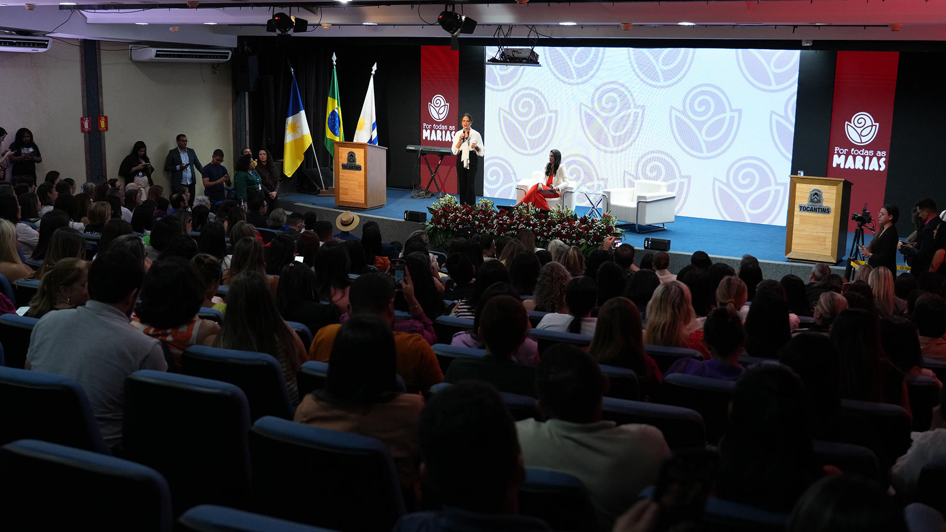 A imagem mostra o auditório lotado, com pessoas sentadas em fileiras assistindo a uma palestra no palco, durante o lançamento da edição 2025 do projeto Por Todas as Marias.