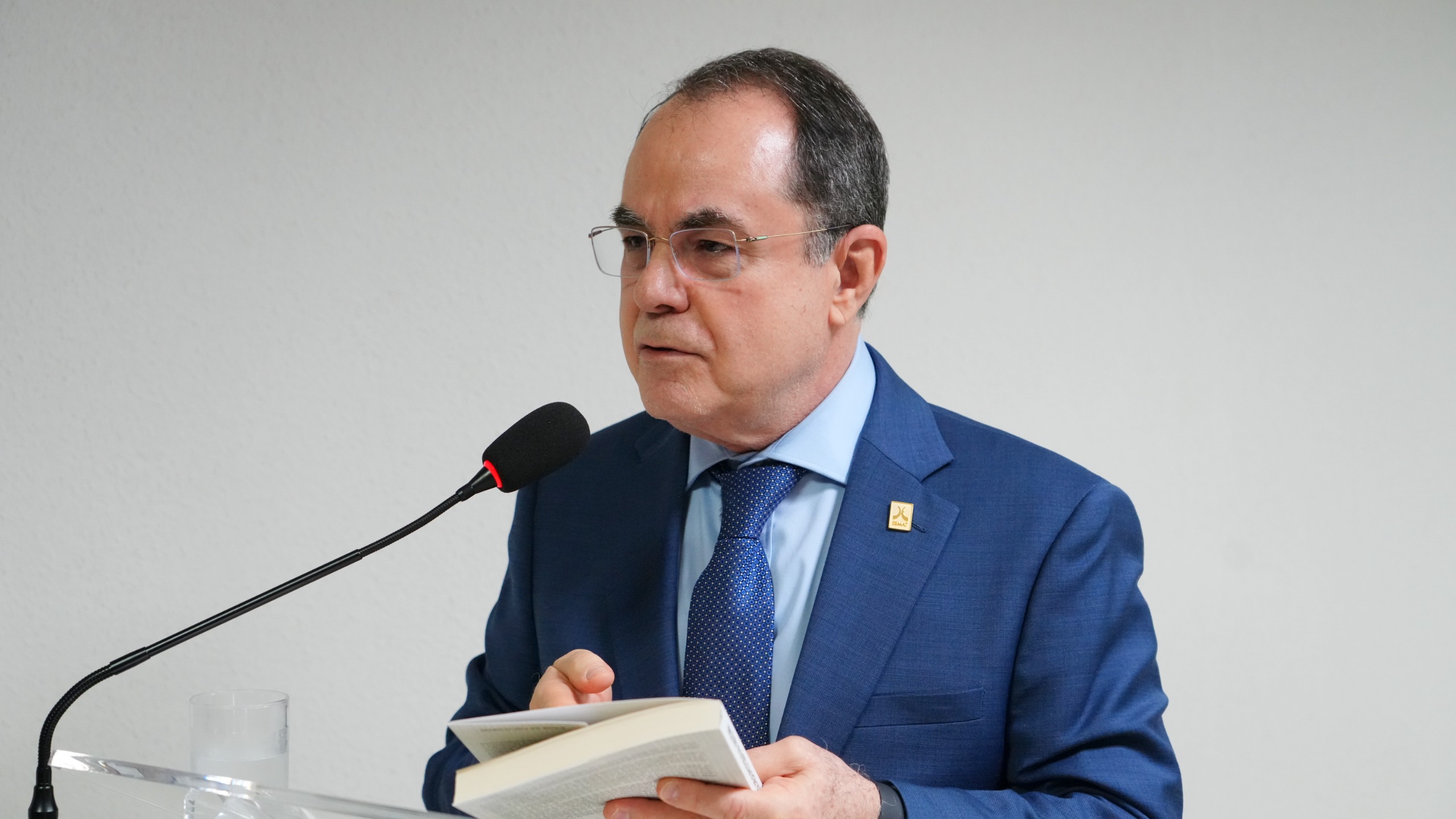 Na imagem, um homem está em pé, falando ao microfone. Ele veste terno azul, camisa azul-clara e gravata azul com pequenos pontos brancos. Usa óculos de grau e segura um livro aberto com a mão esquerda, enquanto parece ler ou citar um trecho. No paletó, há um pequeno broche dourado na lapela. O fundo é liso e claro, transmitindo um ambiente formal, possivelmente institucional ou acadêmico.