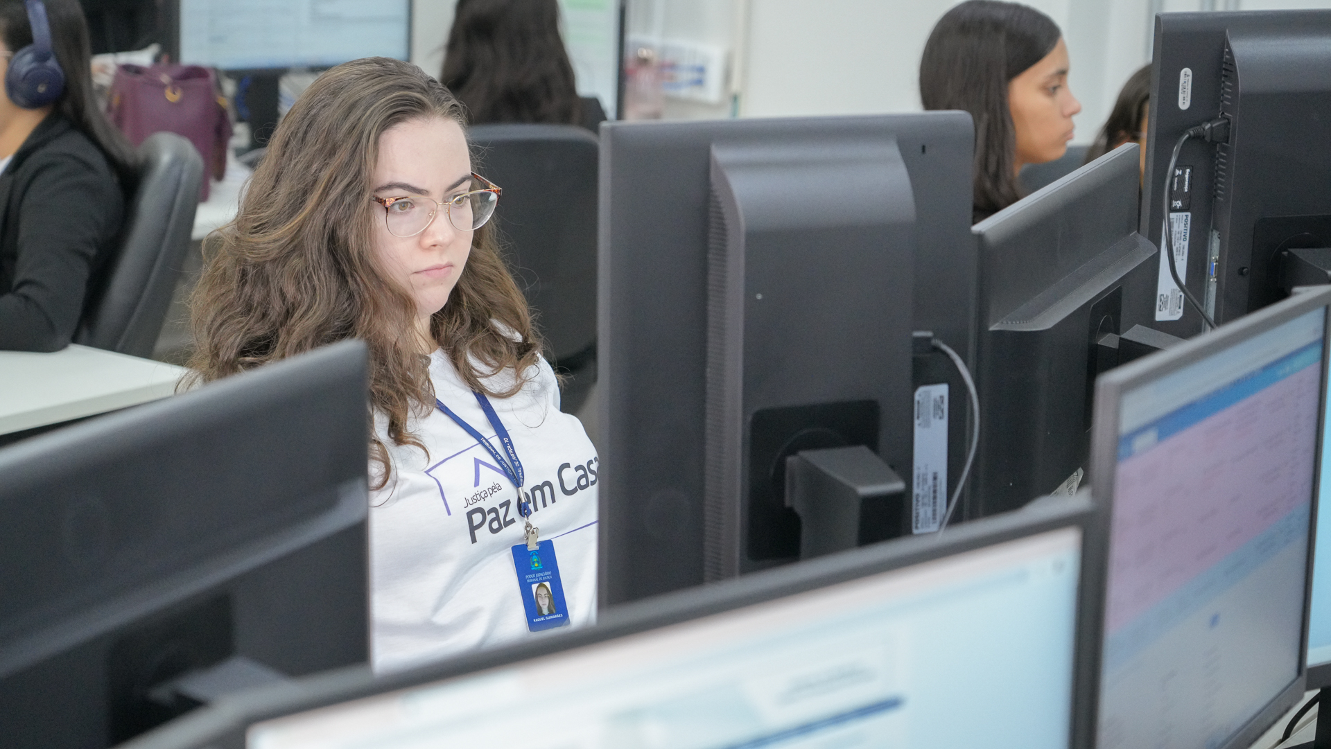 Foto fechada em que aparece uma servidora com camiseta branca da Semana Justiça Pela Paz em Casa e crachá trabalhando no computador; em volta, vários monitores e alguns servidores tranalhando