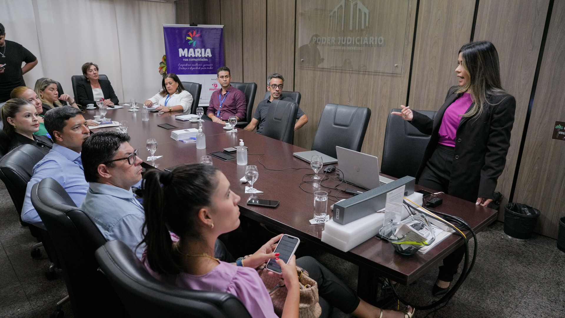 Reunião em uma sala do Poder Judiciário, com cerca de dez participantes sentados ao redor de uma mesa oval, atentos à apresentação de uma mulher em pé à direita da imagem. Ela veste blazer preto e blusa roxa e gesticula enquanto fala. Sobre a mesa, notebooks, copos de água, frascos de álcool em gel e documentos. O ambiente é corporativo e formal, com painel de madeira e o brasão do Poder Judiciário ao fundo.