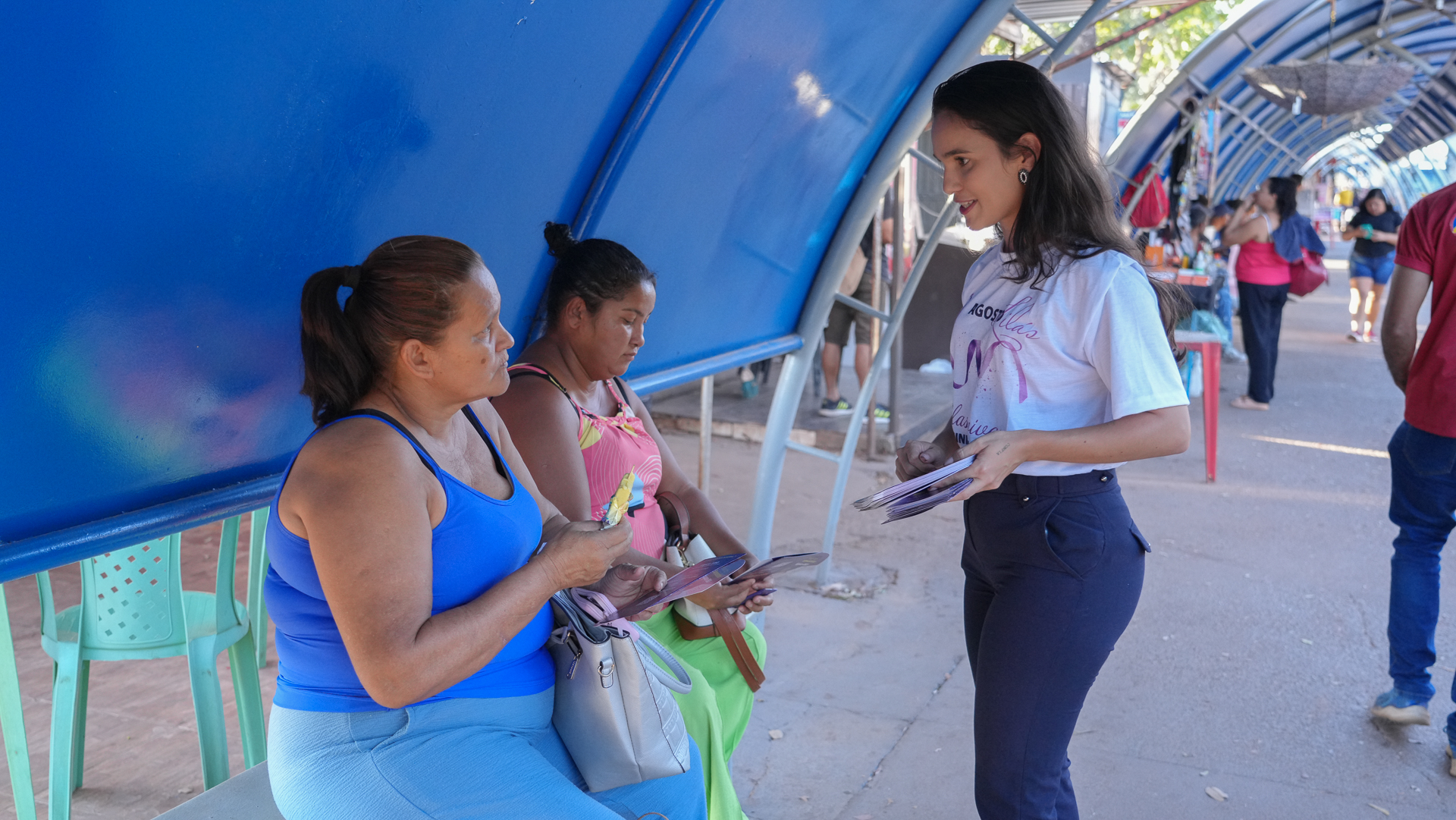 Imagem mostra três mulheres, duas estão sentadas sob um abrigo azul, enquanto a outra mulher, em pé, interage com elas.