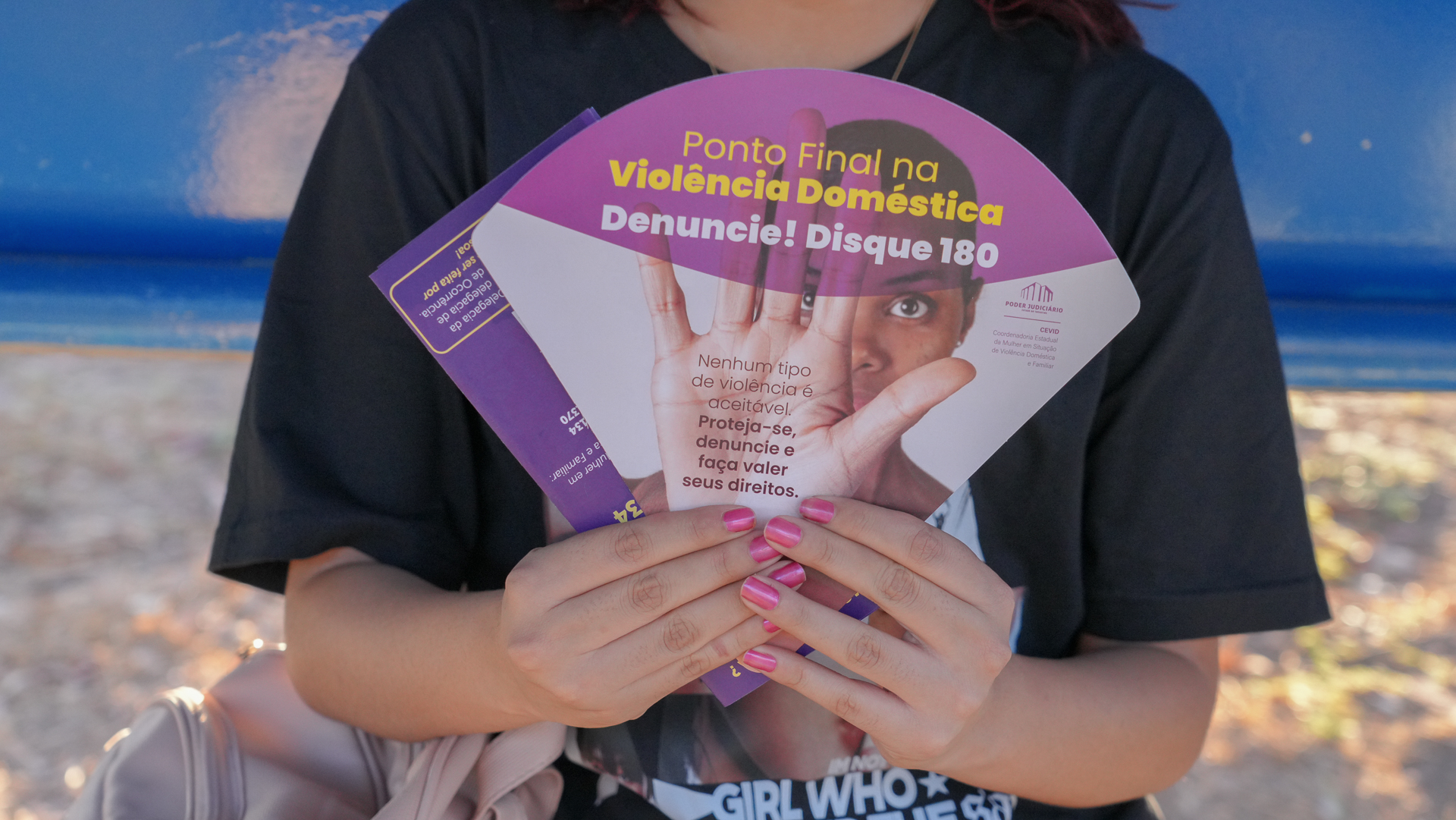  imagem mostra uma pessoa segurando vários panfletos de cor lilás, com o texto em português sobre violência doméstica. 