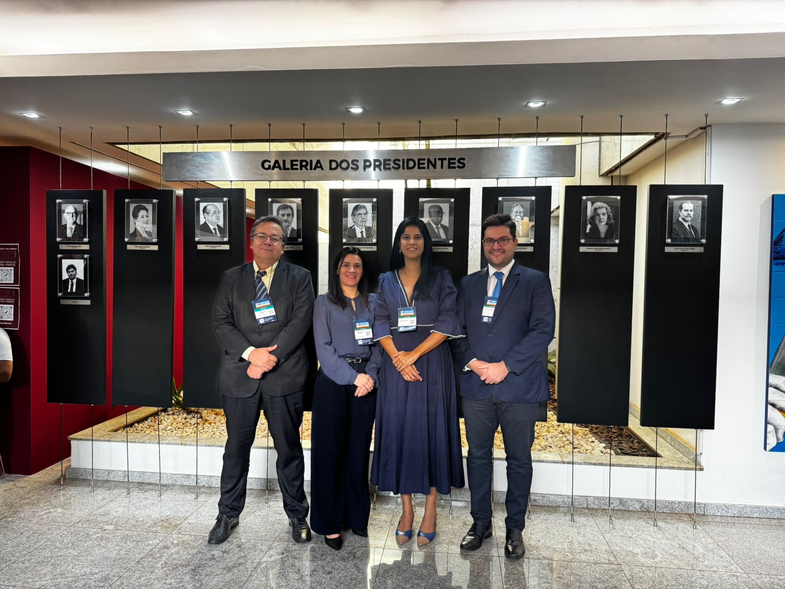 Foto de quatro pessoas, dois homens e duas mulheres ao centro, representando o TJTO no evento; todos estão em pé, posando para a foto, usando roups em tons de cinza e escuros; ao fundo uma galeria de fotos.