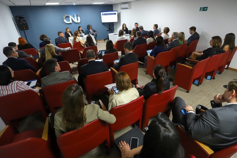Imagem de uma ampla sala com várias pessoas sentadas em cadeiras vermelhas participando de uma reunião 