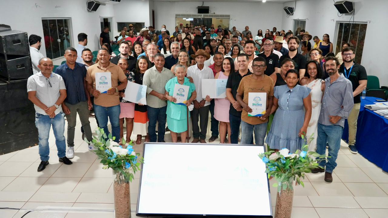 A imagem mostra um grande grupo de pessoas reunidas em uma sala para a cerimônia de entrega de títulos de propriedade em São Félix. A maioria está em pé, organizada em fileiras, algumas sorrindo e outras com expressão neutra, registrando o momento de conquista.