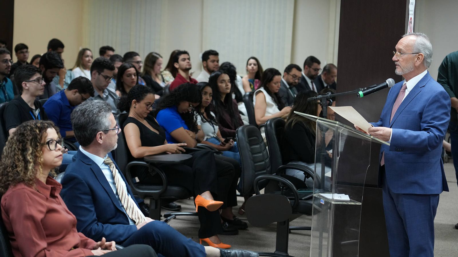 A imagem mostra o salão do Júri durante a Correição Geral Ordinária na Comarca de Palmas, com várias pessoas sentadas em cadeiras prestando atenção, enquanto um homem fala no púlpito.