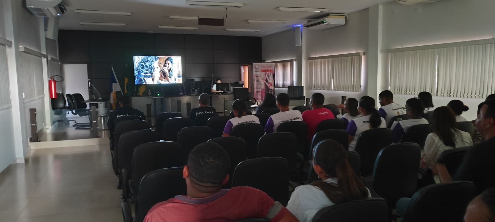 Uma sala grande com um grupo de pessoas sentadas, assistindo um filme na tela.