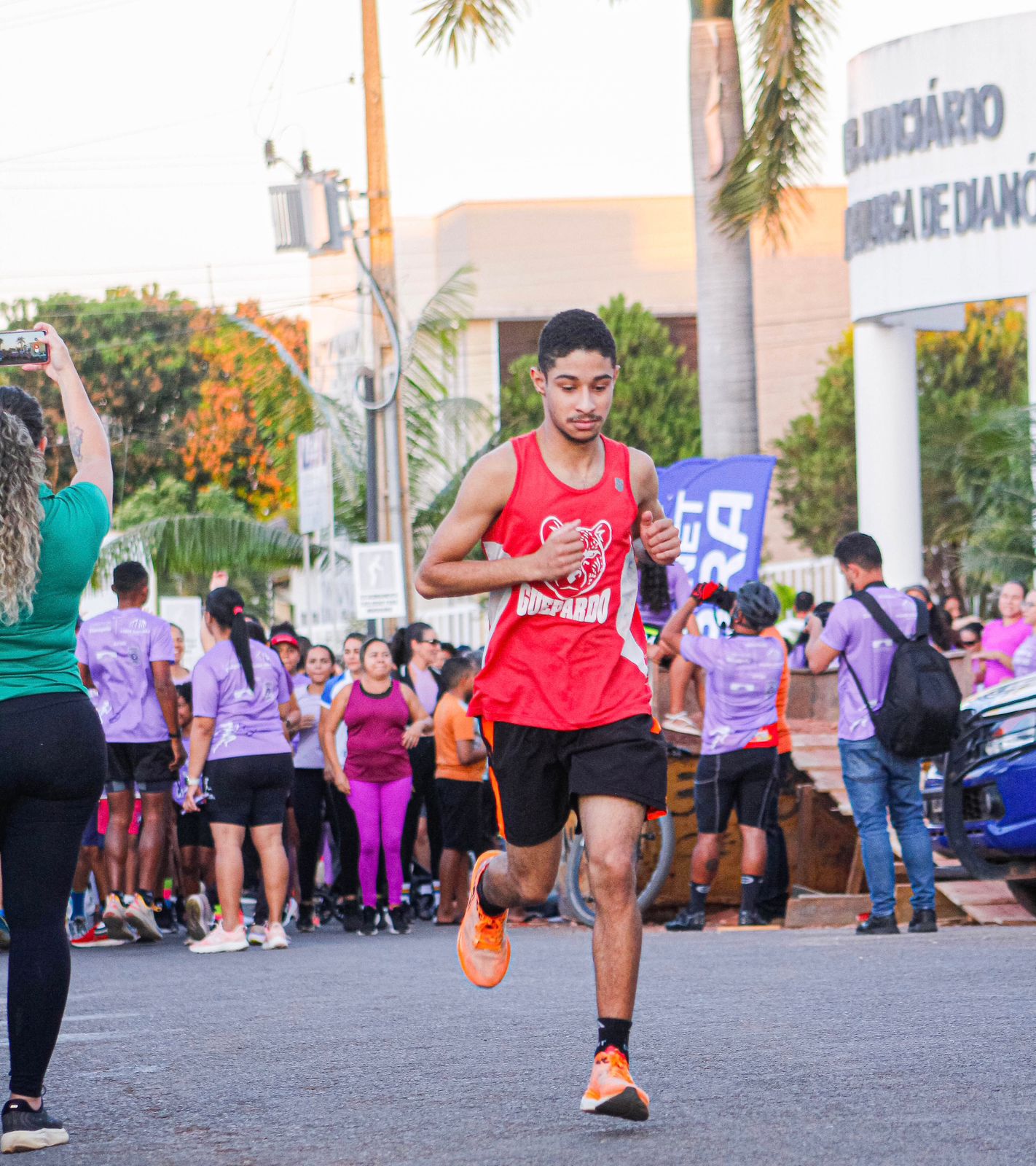 Imahm mostra um corredor com bermuda preta e camiseta vermelha em primeiro plano e ao fundo um grande grupo de pessoas concentradas em frente ao Fórum de Dianópolis para participação na corrida