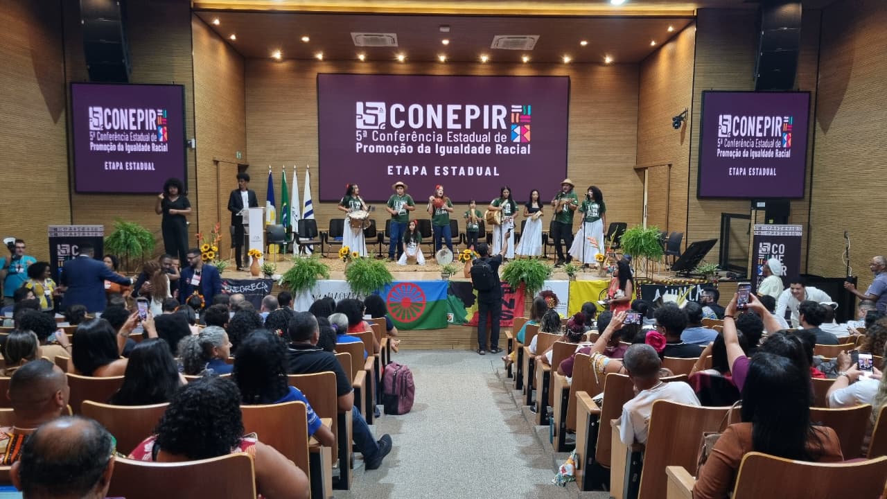 A imagem mostra o auditório do Tribunal de Justiça do Tocantins durante a 5ª Conferência Estadual de Promoção da Igualdade Racial. O auditório está lotado de pessoas sentadas e, no palco, ocorre uma apresentação cultural.