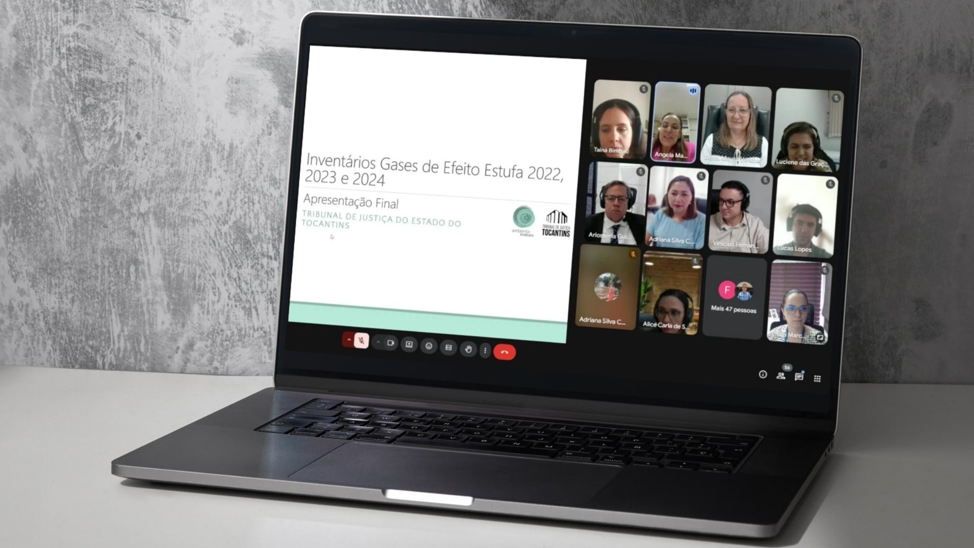 Foto de um notebook sobre uma mesa clara, com a tela aberta em uma videoconferência. À esquerda, está projetado um slide com o título: “Inventários Gases de Efeito Estufa 2022, 2023 e 2024 – Apresentação Final”, acompanhado dos logotipos do Instituto Ambienta e do Tribunal de Justiça do Estado do Tocantins. À direita, aparecem várias janelas da reunião on-line, com rostos de participantes — homens e mulheres — alguns usando fones de ouvido.