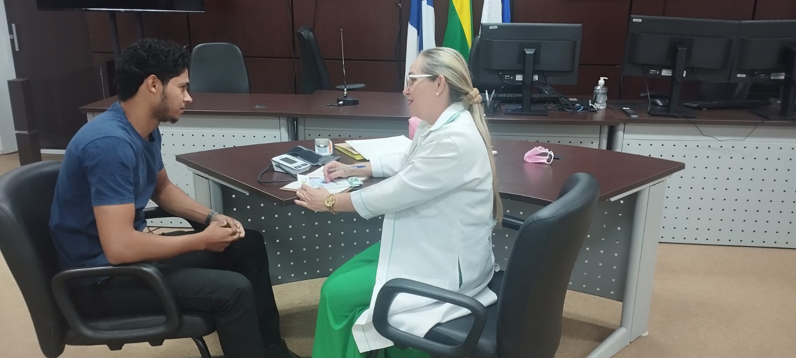 Foto aberta de uma técnica da área de saúde fazenda atendimento a um servidor; ambos estão sentados de frente, ao lado de uma mesa, sendo que a técnica está do lado direito e o servidor à esquerda