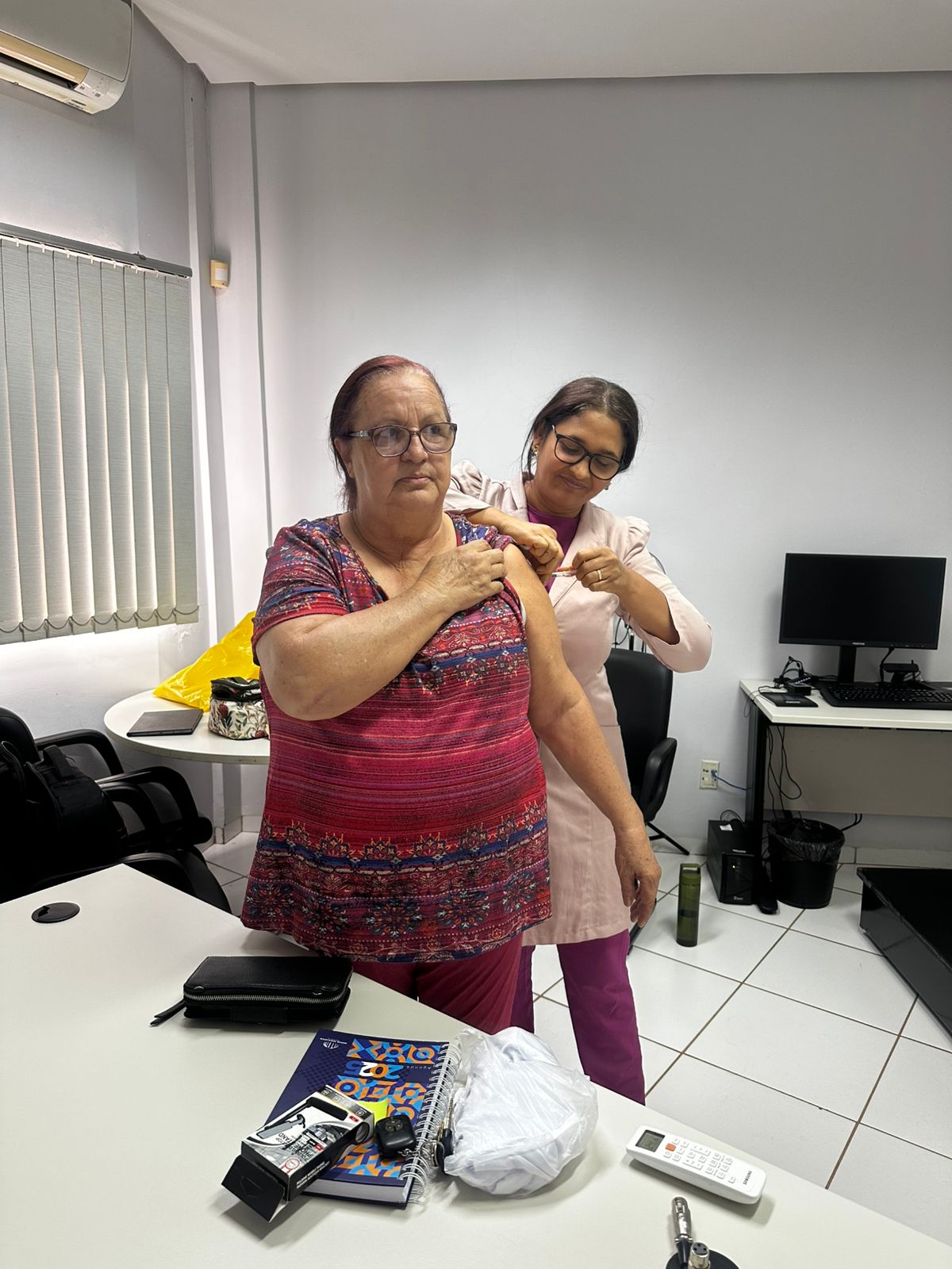 Em uma sala, servidora está de frente, recebendo vacina aplicada por uma técnica de saúde, que também está de frente. 
