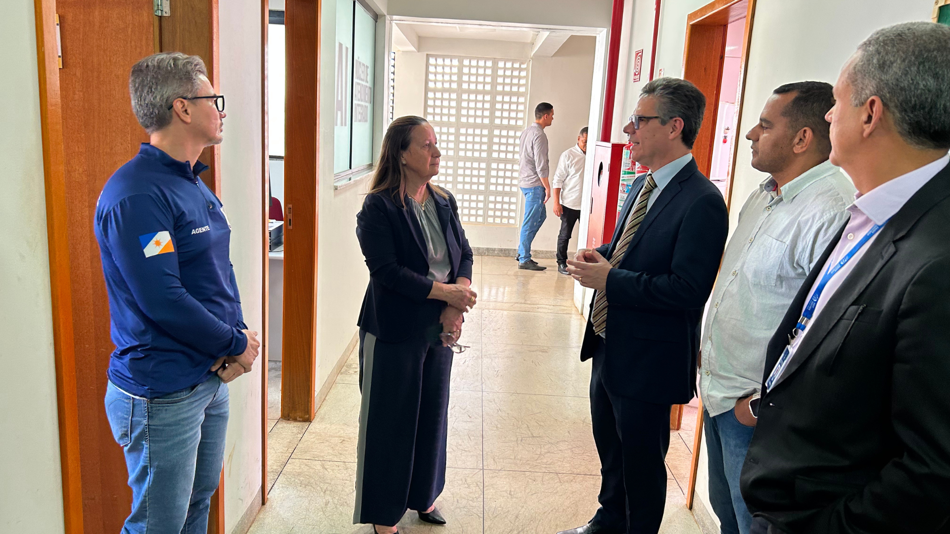 Em um corredor de paredes claras e portas de madeira, cinco pessoas estão em pé, conversando. Ao centro, uma mulher de cabelos castanhos claros presos, veste blazer preto e calça escura, dialoga com um homem de terno preto, camisa clara e gravata listrada, que gesticula com as mãos. À esquerda, um agente de uniforme azul marinho, com o brasão do Tocantins no braço, observa a conversa. À direita, dois homens acompanham: um de camisa branca e outro de terno escuro com crachá azul. Ao fundo, próximo a uma parede de blocos de vidro que deixa a luz natural entrar, duas pessoas caminham pelo corredor.