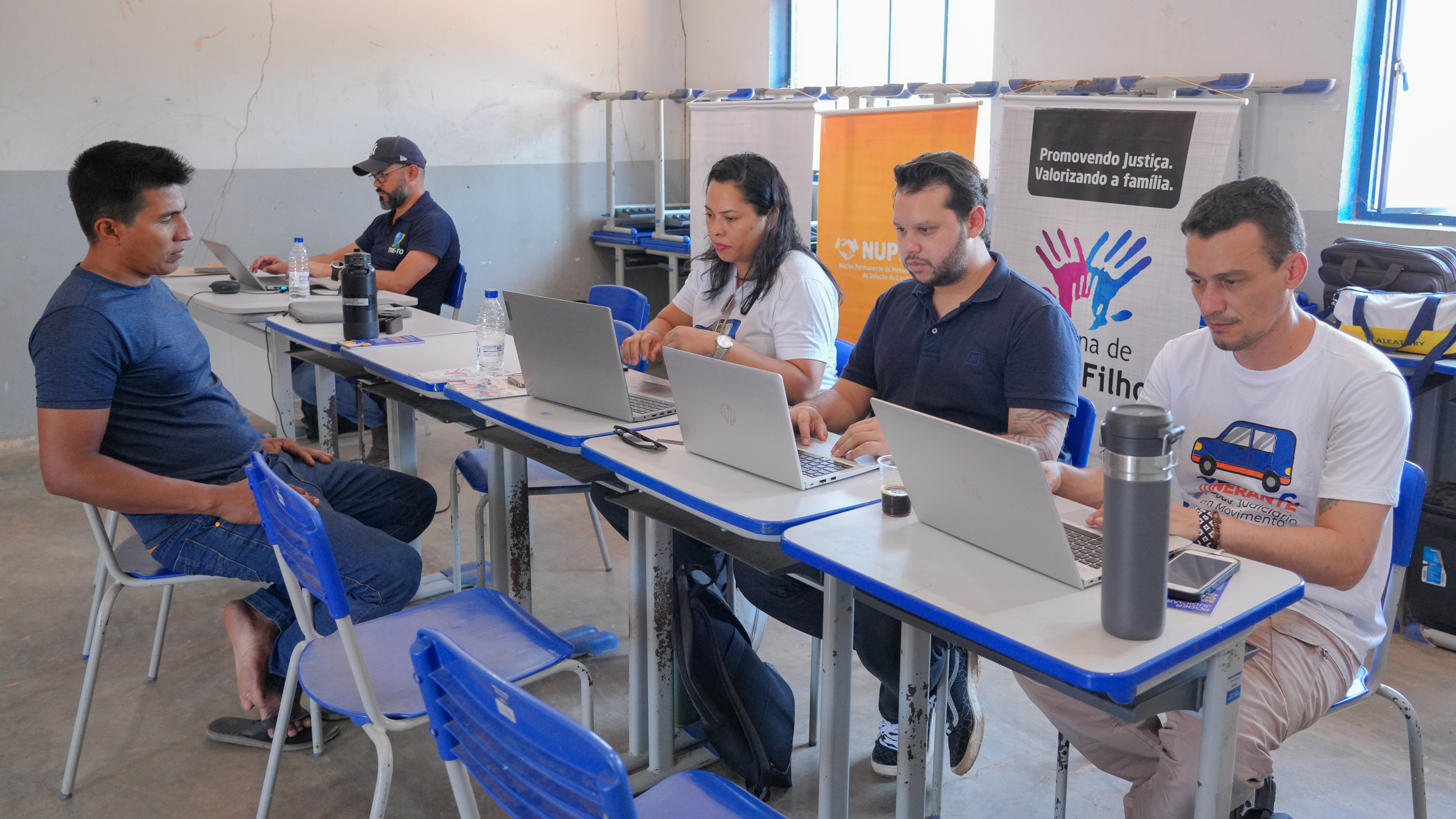 Imagem mostra vários pessoas sentadas em torno de mesas, trabalhando em computadores portáteis em uma sala. Uma das pessoas atende um indígena.