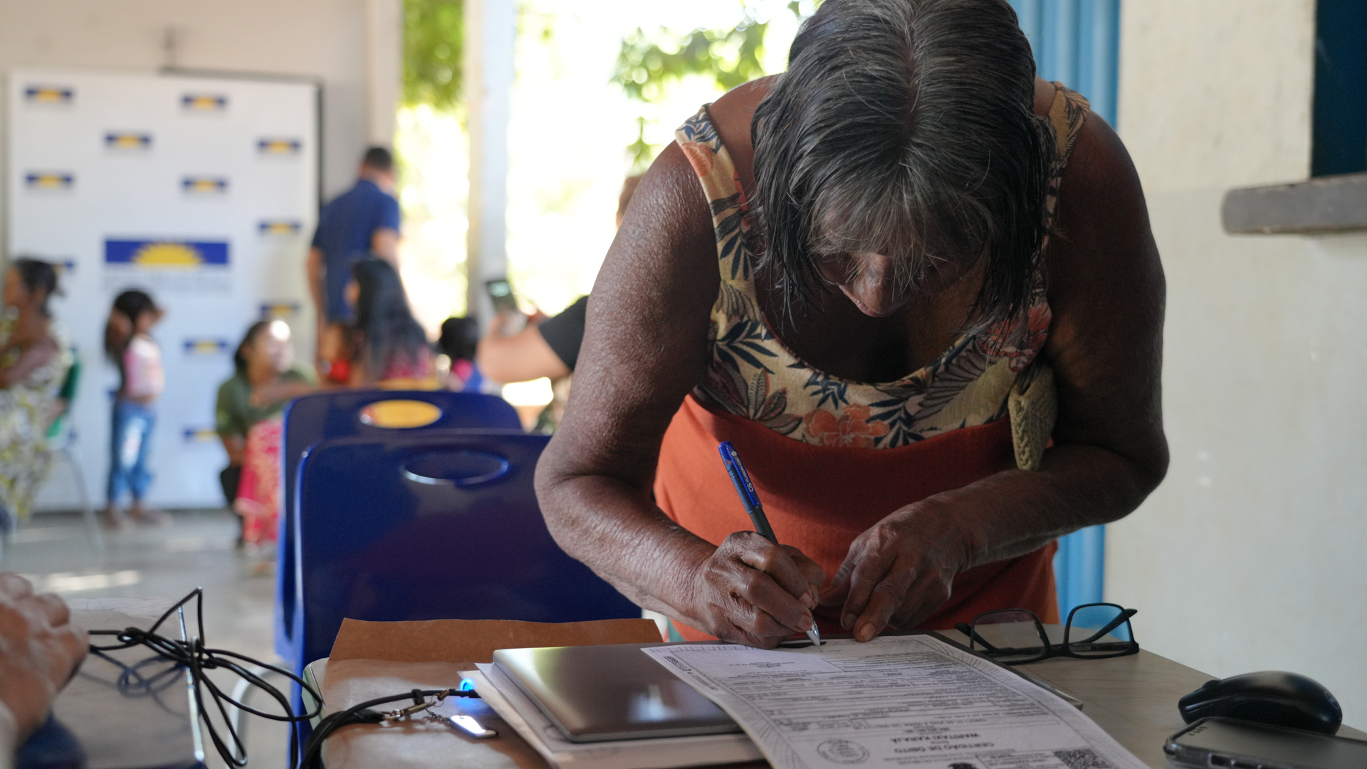 Mulher indígena de meia-idade, inclinada sobre uma mesa, assinando um documento.
