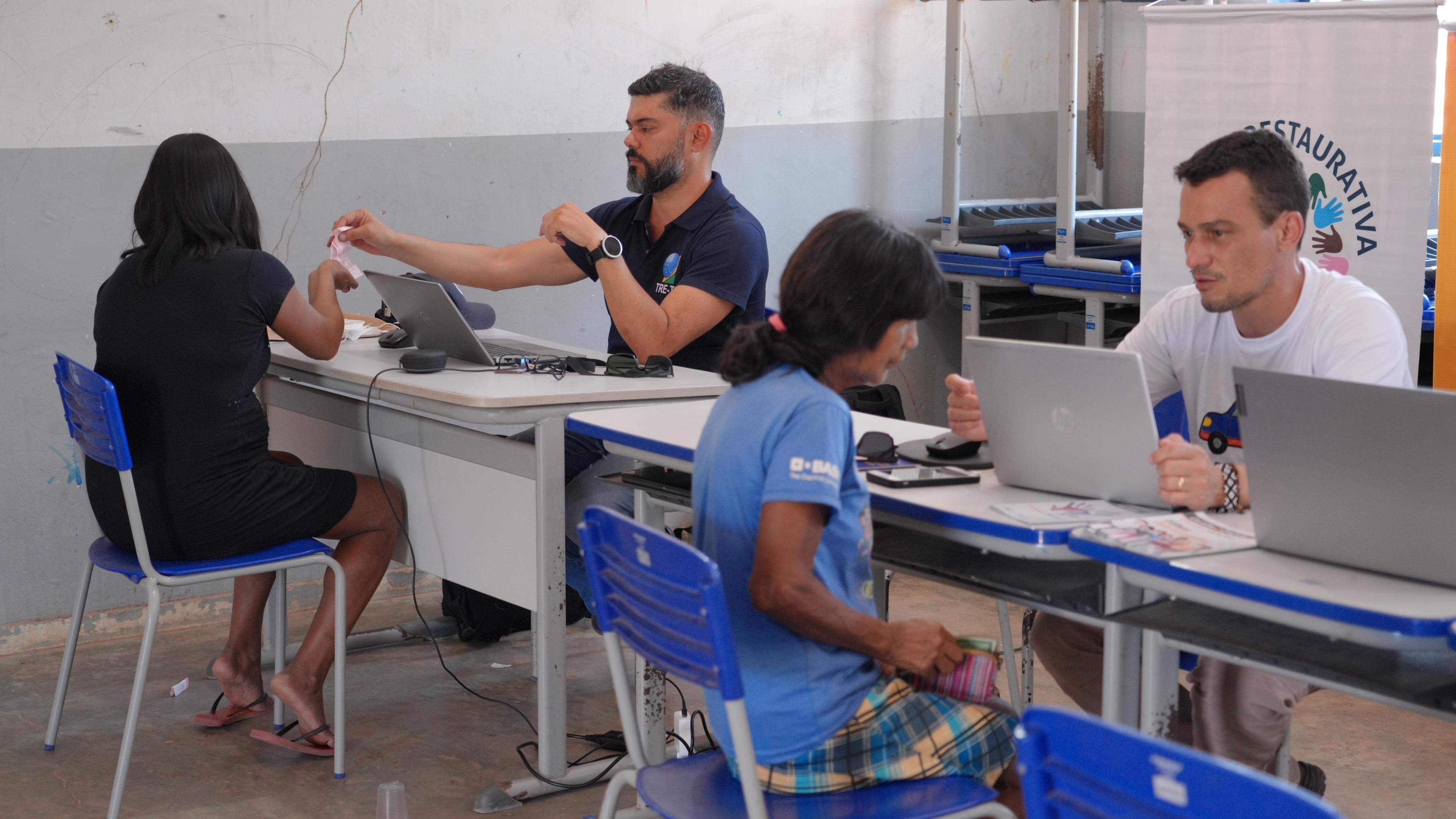 Imagem mostra pessoas sentadas em volta de mesas com computadores, em uma sala, prestando atendimento a indígenas