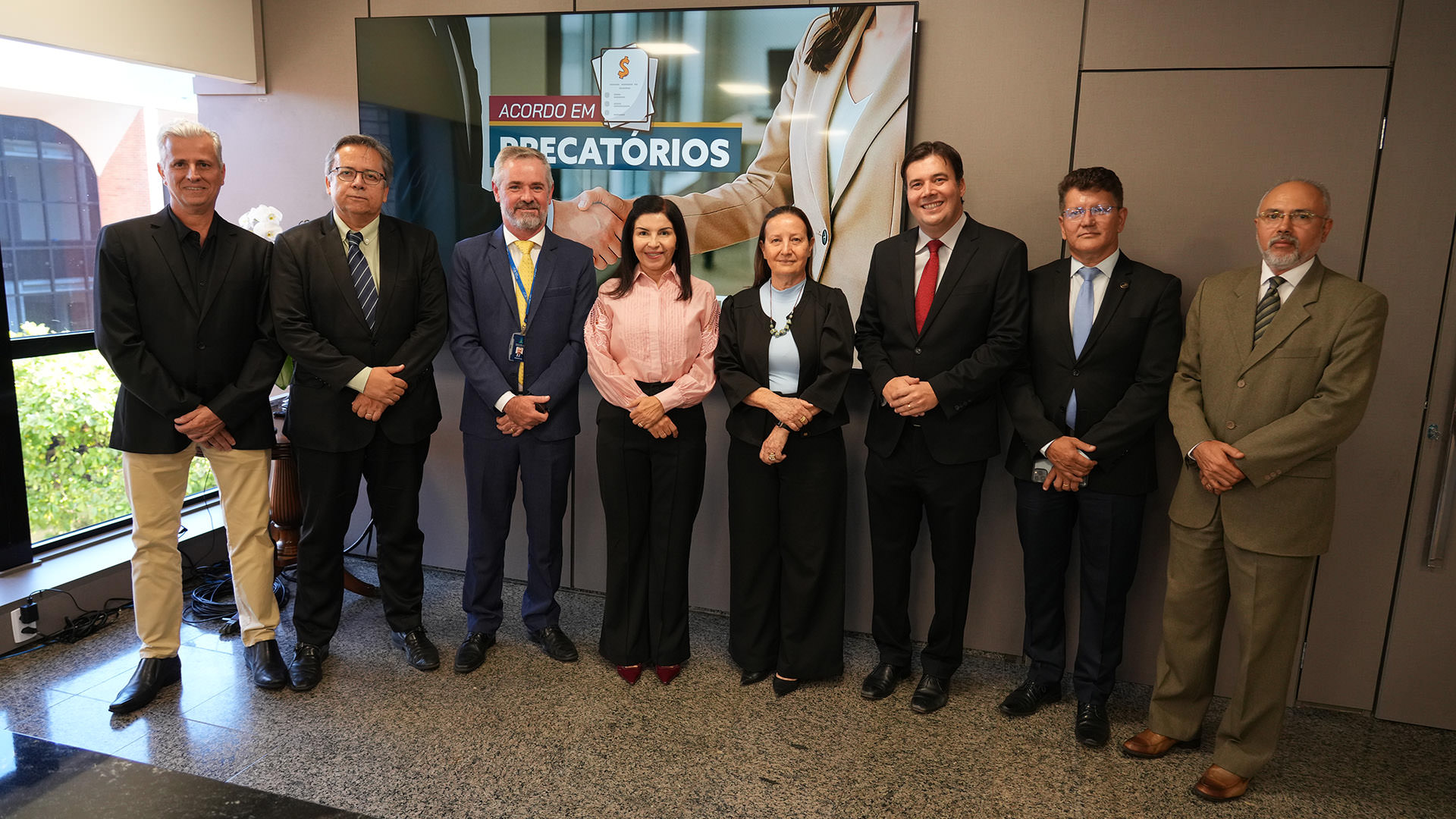 A imagem mostra oito pessoas estão alinhadas e posam para a foto em ambiente interno, diante de uma tela que exibe a frase “Acordo em Precatórios”. Entre elas, homens de terno e mulheres em trajes sociais, todos sorridentes e olhando para a câmera.