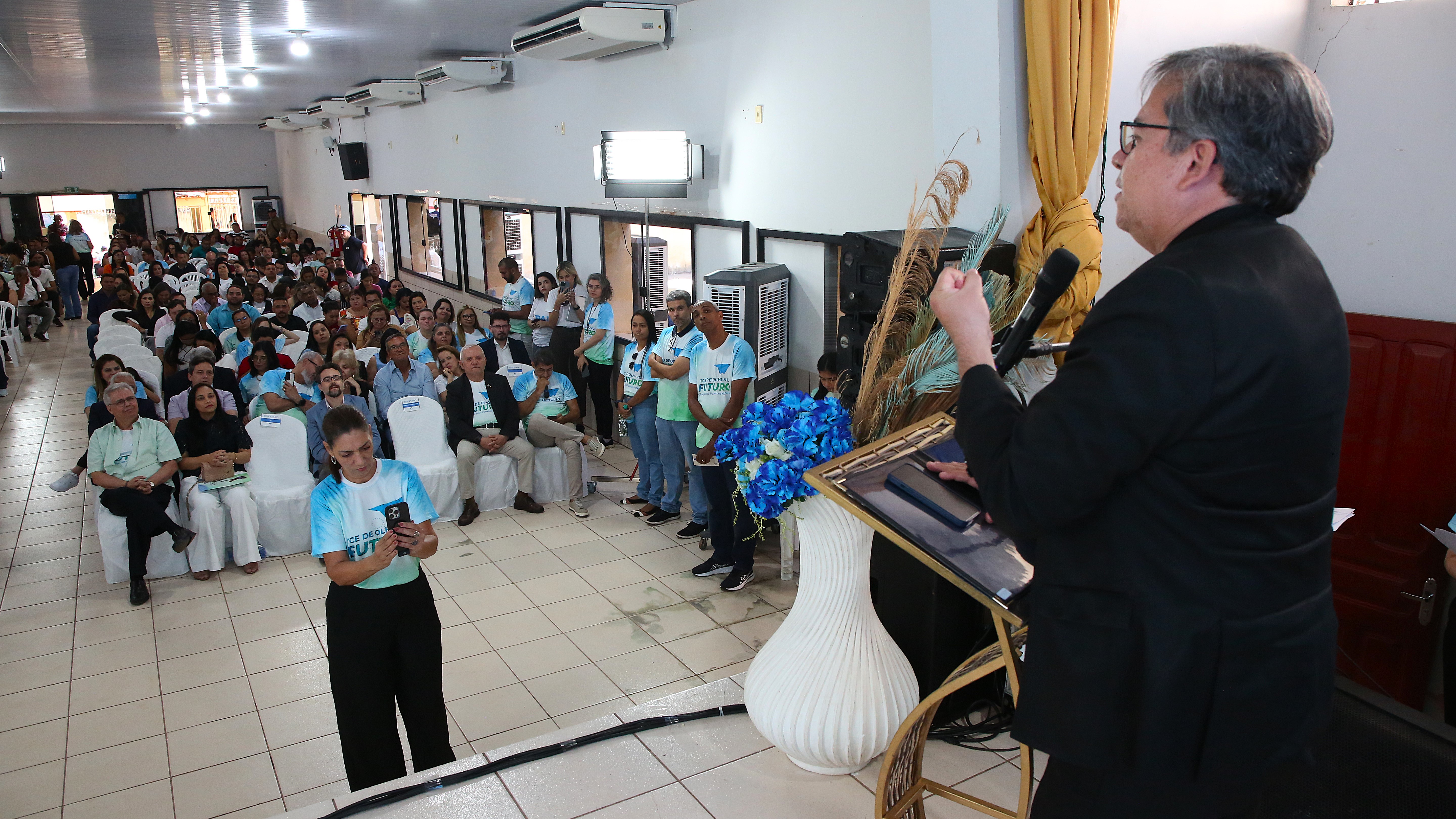Homem pardo de cabelos grisalhos e terno preto fala de costas ao púlpito para plateia em auditório de paredes brancas