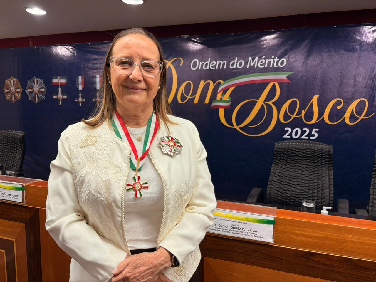 A presidente do TJTO posa sorridente após receber a Medalha da Ordem do Mérito Dom Bosco 2025. Ela veste um conjunto claro, com bordados no blazer, e está com medalha em fita verde, branca e vermelha e uma insígnia da Ordem presa à roupa. Ao fundo, o painel azul com a inscrição “Ordem do Mérito Dom Bosco 2025” e detalhes dourados.