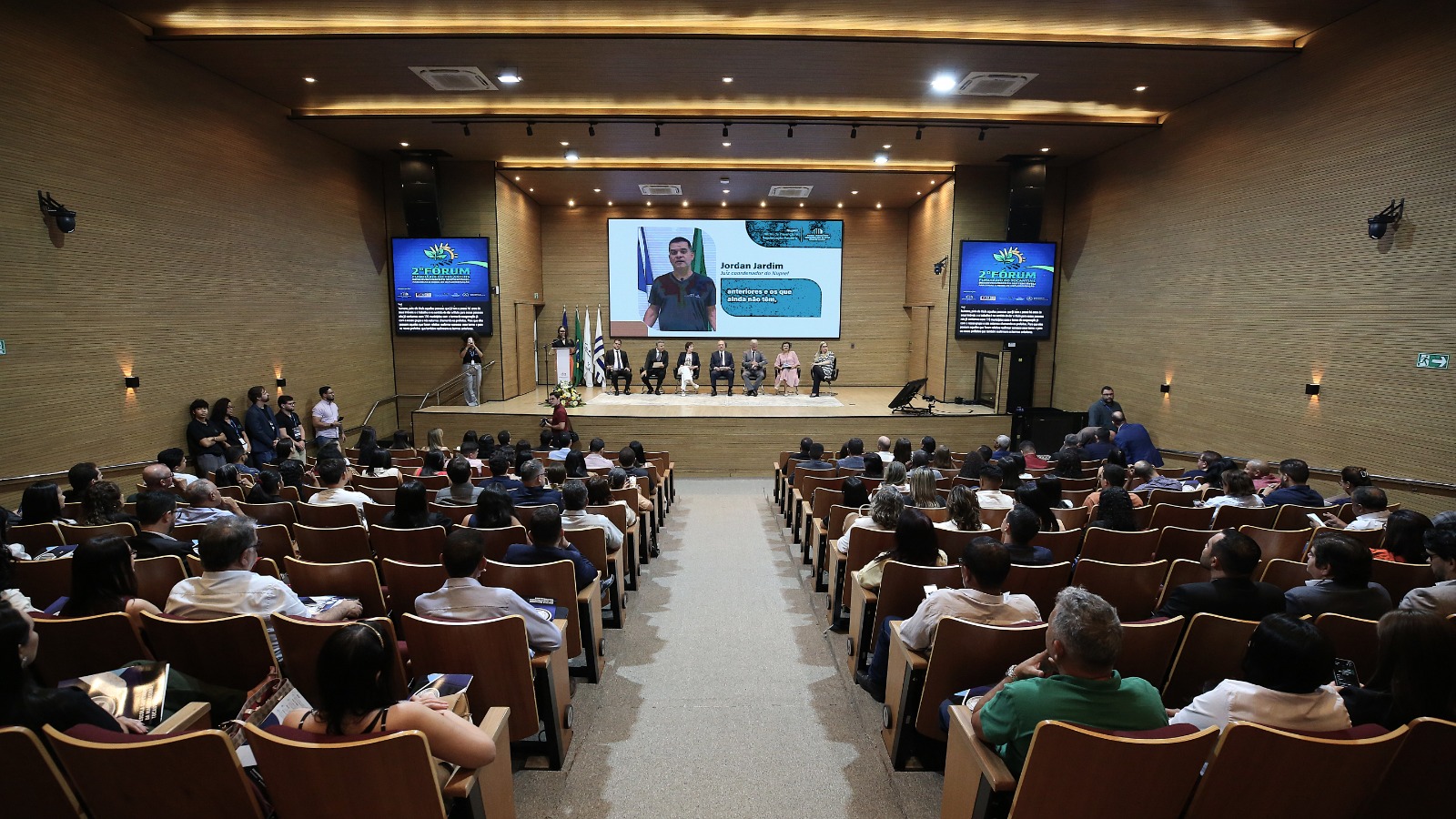 Homens e mulheres sentados em auditório lotado assistem palestra em telão ao fundo de palco. O espaço tem paredes e teto em acabamento em madeira