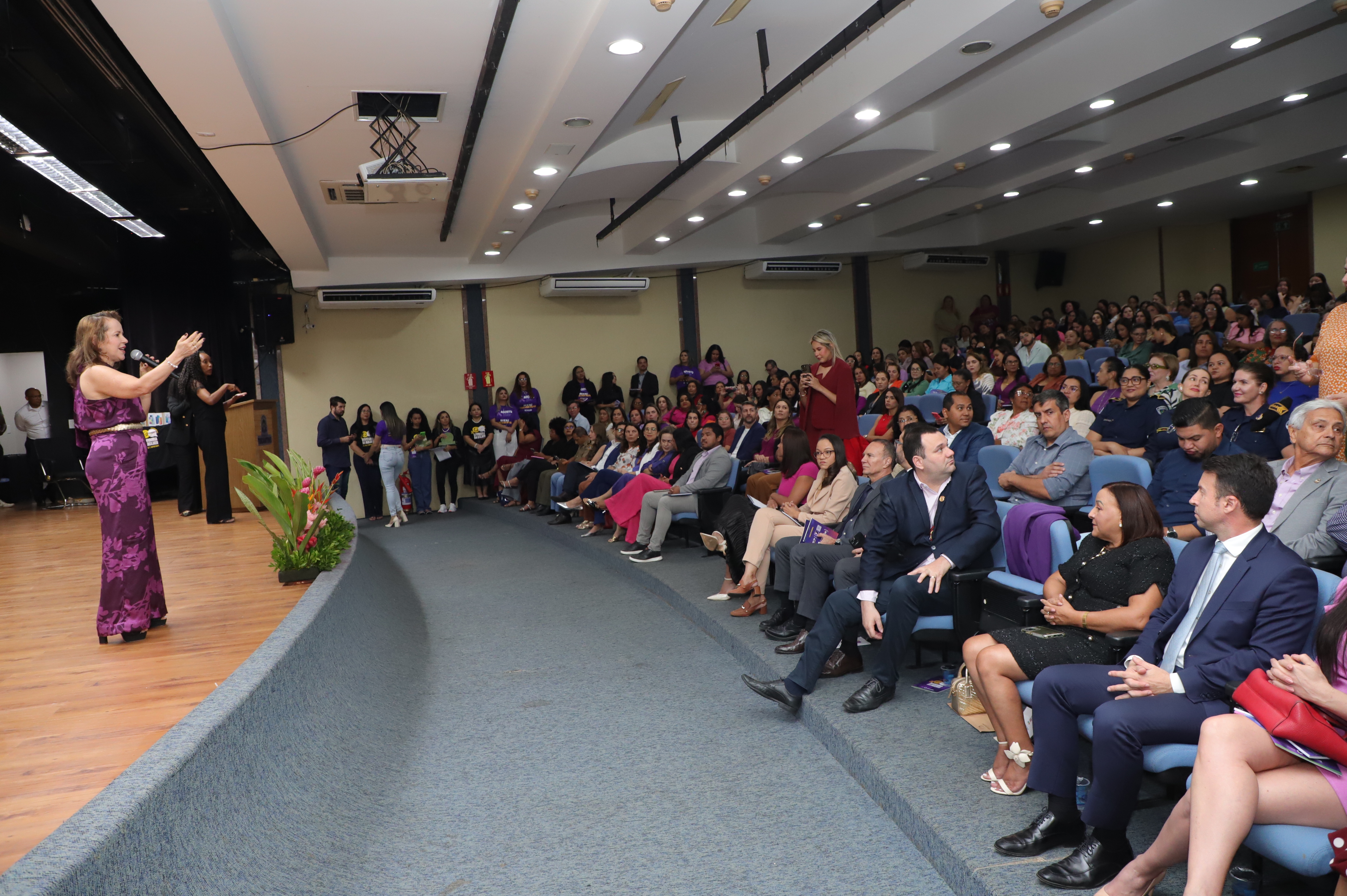 Foto aberta com a juíza falando ao microfone à esquerda, no palco do auditório; e à direita o público assiste.