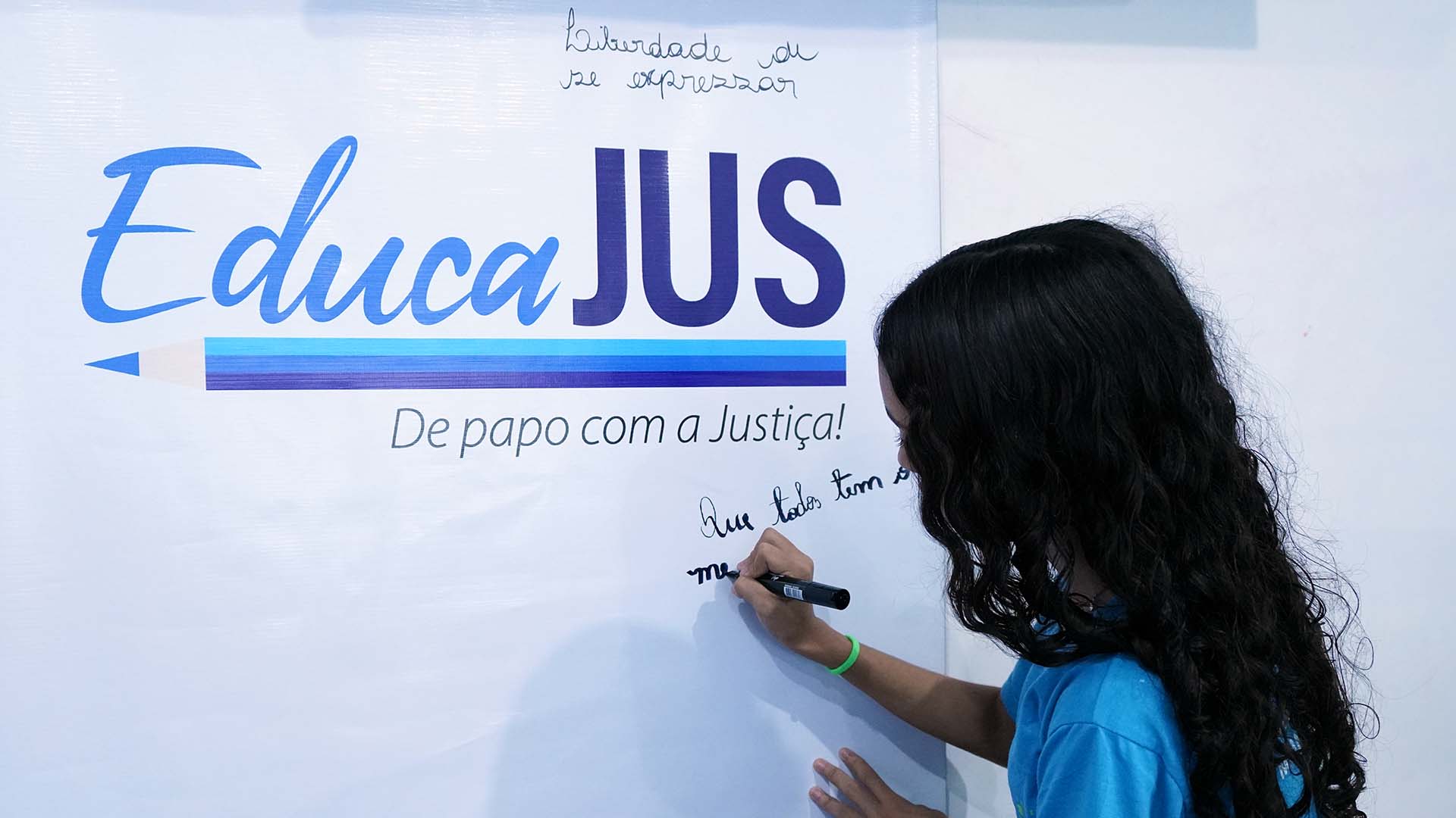 Menina de cabelos longos cacheados escrevendo em lousa branca que apresenta logo do programa EducaJus