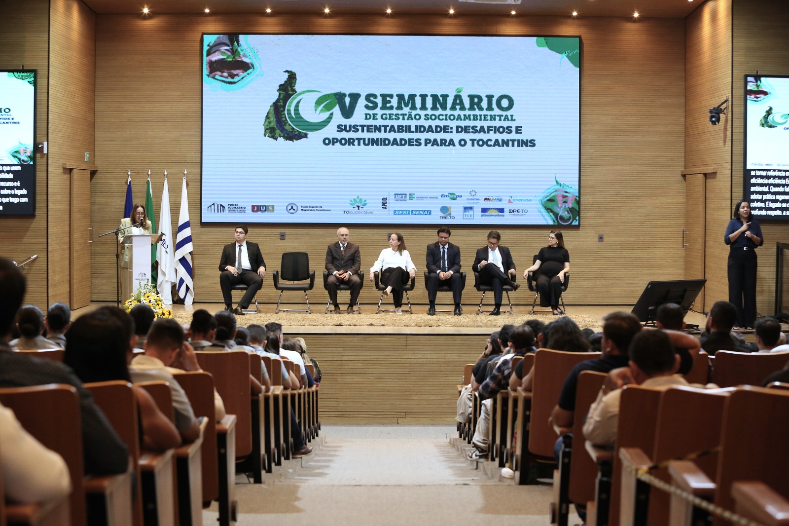 Imagem aberta de um auditório cheio de pessoas assistindo a uma palestra. No palco, sete cadeiras com seis pessoas sentadas e uma mulher falando no microfone ao púlpito