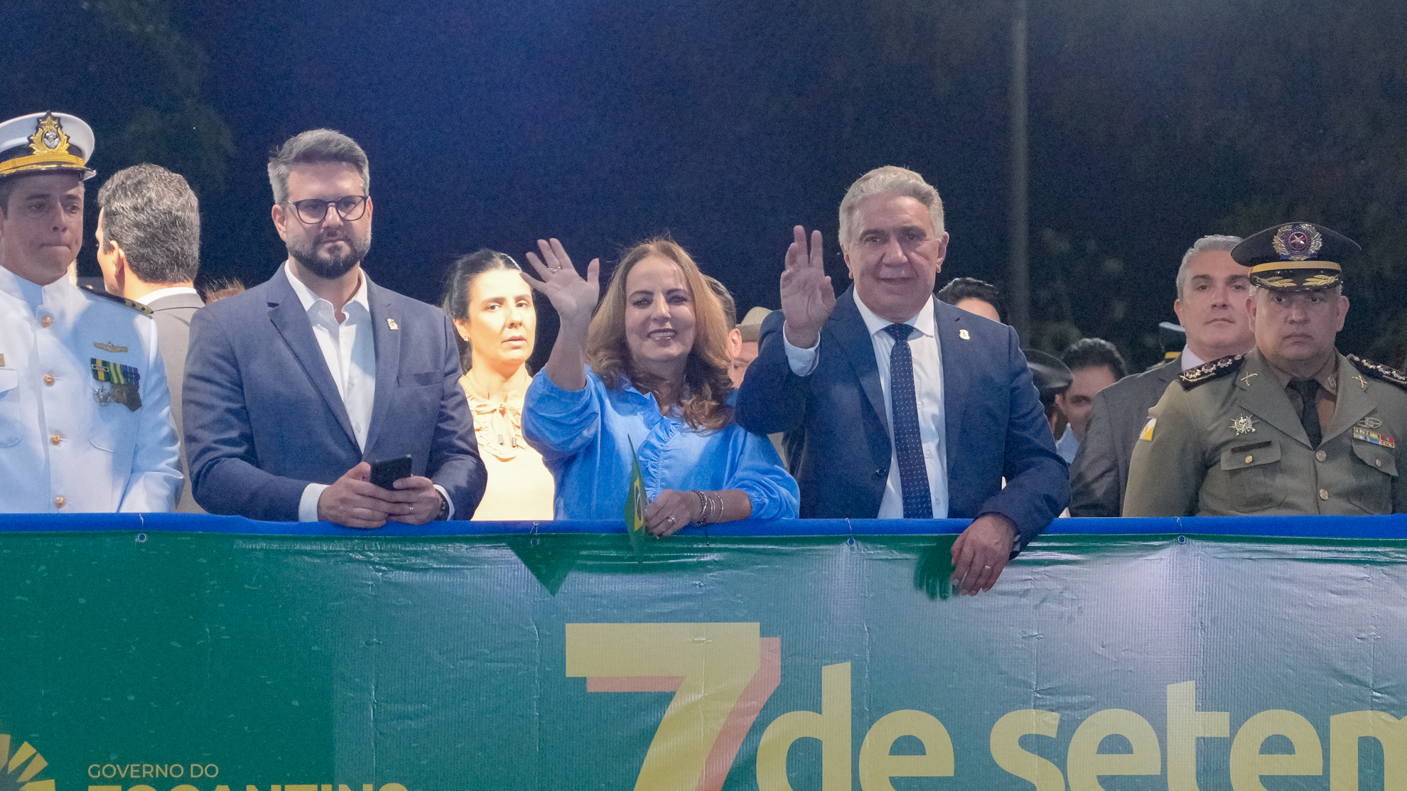Nas imagens aparece um palanque oficial das comemorações de 7 de Setembro, à noite, há várias autoridades lado a lado. No centro, uma mulher de blusa azul sorri e acena para o público. À sua direita, um homem de terno escuro e gravata azul também acena. Eles estão rodeados por outras autoridades civis e militares: à esquerda, um oficial da Marinha em uniforme branco; à direita, um militar do Exército em uniforme verde-oliva com boina. À frente do grupo, uma faixa verde traz a inscrição “7 de setembro” acompanhada da logomarca do Governo do Tocantins. O ambiente é iluminado artificialmente e transmite solenidade.
