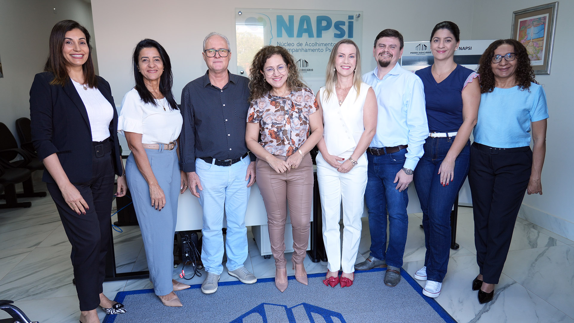 Imagem de um grupo de profissionais posicionado lado a lado, em fileira, sendo seis mulheres e dois homens; ao fundo uma placa transparente com o nome NAPsi em azul
