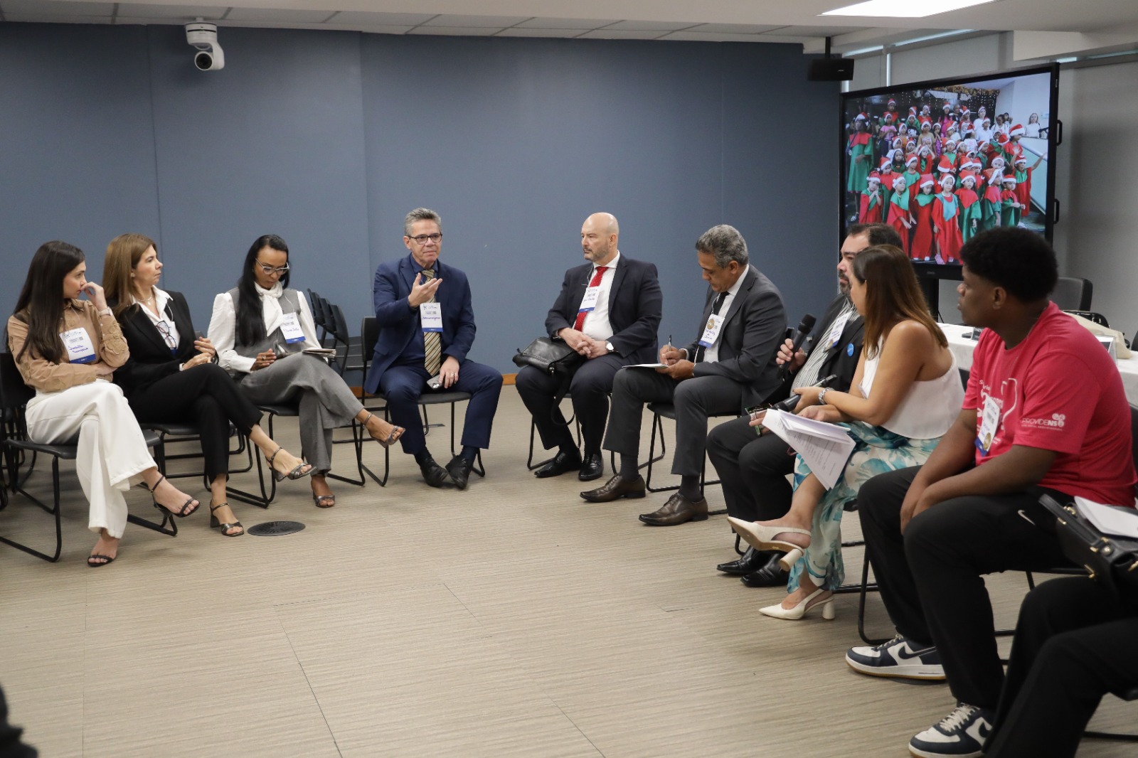 Um grupo de dez pessoas participa de uma roda de conversa em uma sala de reuniões. Elas estão sentadas em cadeiras organizadas em círculo. A maioria veste roupas formais, como ternos, blazers e calças sociais, enquanto duas pessoas à direita usam trajes mais informais: uma mulher com vestido estampado azul e branco e um jovem com camiseta vermelha e calça preta. No centro do grupo, um homem de terno azul fala, gesticulando com a mão, enquanto outros escutam atentamente, fazem anotações ou seguram microfones e papéis.