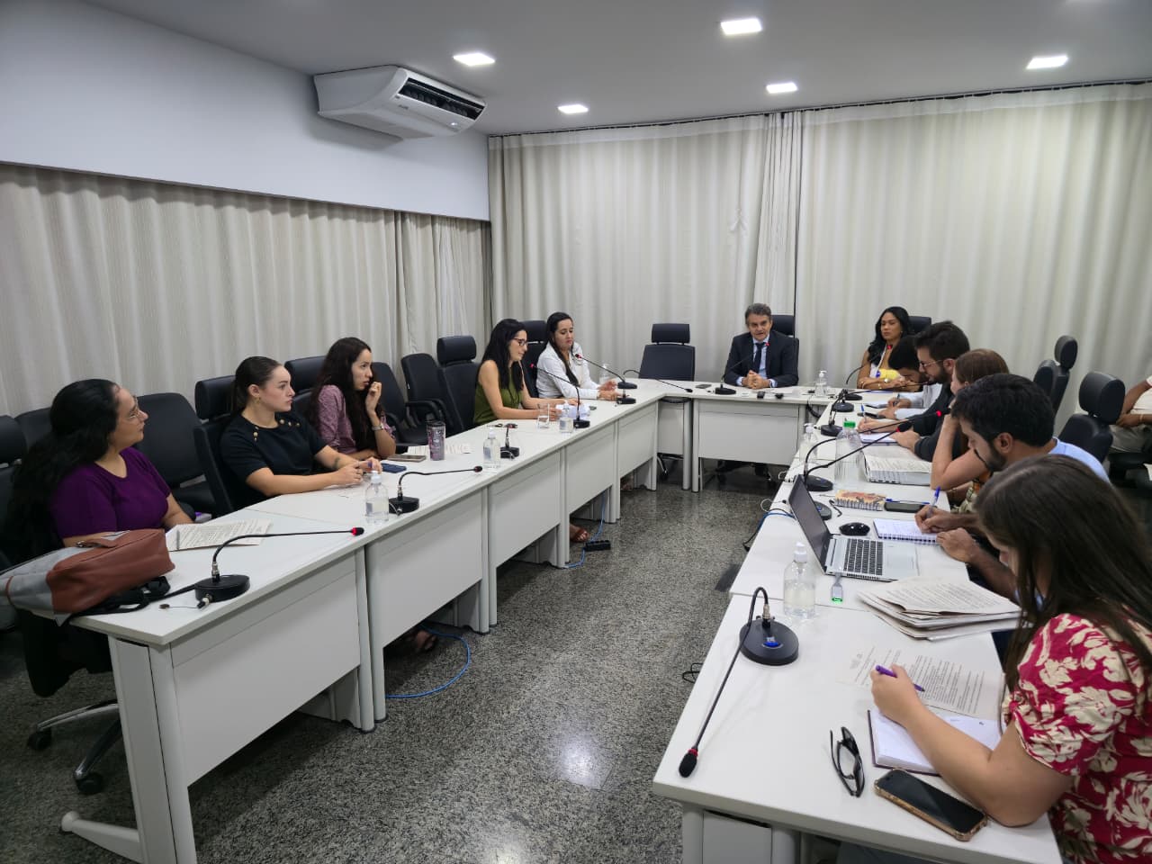 Homens e mulheres sentados em mesa branca em formato U, em sala de paredes brancas e persiana bege