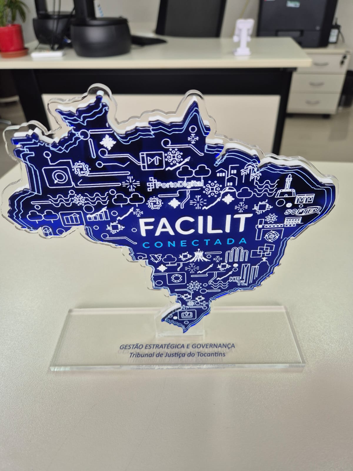 A foto mostra um troféu em acrílico, recortado no formato do mapa do Brasil. Ele é azul, com ícones brancos que remetem a tecnologia, inovação e conectividade — como nuvens, gráficos, computadores e símbolos digitais. No centro, em destaque, aparece o nome do evento: "FACILIT CONECTADA".