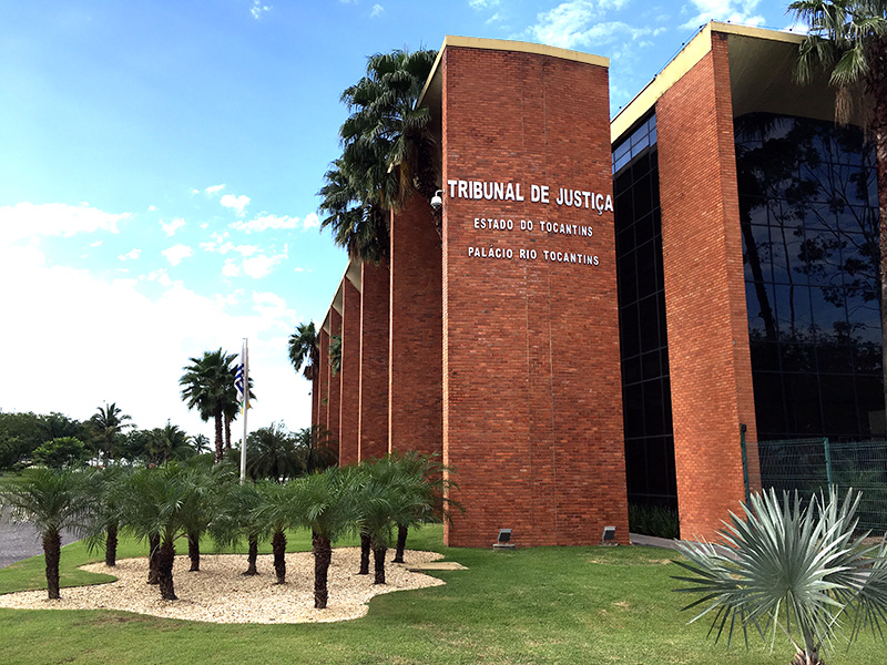 Na imagem, aparece a fachada do prédio do Tribunal de Justiça do Tocantins – Palácio Rio Tocantins, em tijolos avermelhados e janelas de vidro escuro. Em frente, há um jardim com gramado verde, pequenos coqueiros e palmeiras. O céu está azul com algumas nuvens.