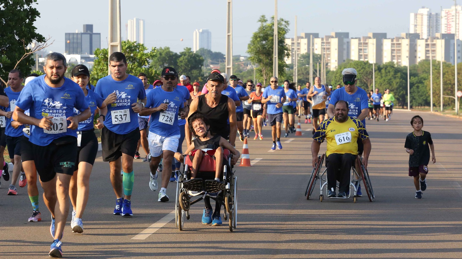 A cena mostra uma corrida de rua inclusiva em uma avenida larga, sob céu claro. Em primeiro plano, vários participantes correm usando camisetas azuis com números de identificação presos ao peito. Entre eles, destacam-se duas pessoas em cadeiras de rodas participando da prova: uma sendo empurrada por um homem de camiseta preta e boné, sorrindo de forma contagiante; a outra, com camiseta amarela, avança de forma determinada, também empurrada por um participante. Ao lado, uma criança corre acompanhando o grupo. Ao fundo, há mais corredores de azul e, ainda mais distante, prédios altos que compõem a paisagem urbana. O clima é de superação, inclusão e celebração esportiva.