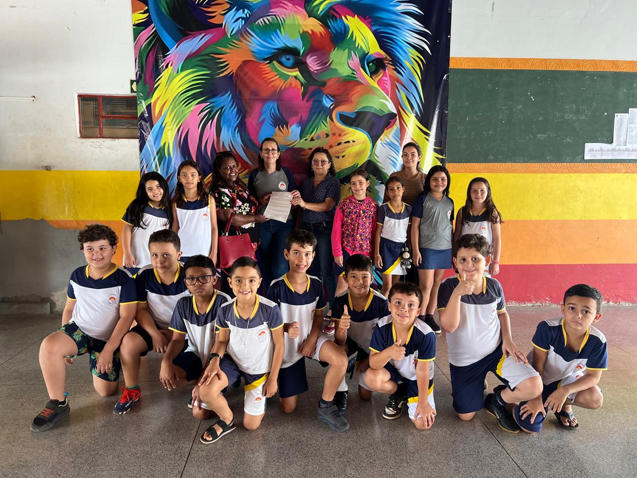 A imagem mostra um grupo de estudantes e três adultos pousando para uma foto e ao fundo tem um quadro de um leão colorido.