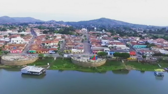 A imagem mostra a orla da cidade de Xambioá. Em primeiro plano está o rio Araguaia, com o flutuante conhecido como “Titanic”. Ao fundo, aparecem várias residências, uma igreja e, logo atrás dela, alguns morros.