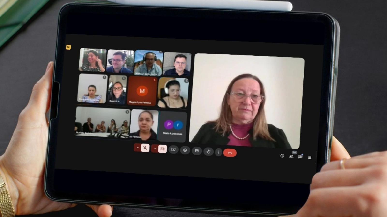 A imagem mostra a primeira reunião virtual com a Comarca de Arapoema para alinhamento do Projeto JUS em Ação. Em destaque, a tela de um tablet sendo segurado exibe a videoconferência. No lado esquerdo, aparece a grade com os participantes, cada um em pequenos quadrados com suas imagens. No lado direito, em tela maior, está a presidente do TJTO, desembargadora Maysa Vendramini Rosal.