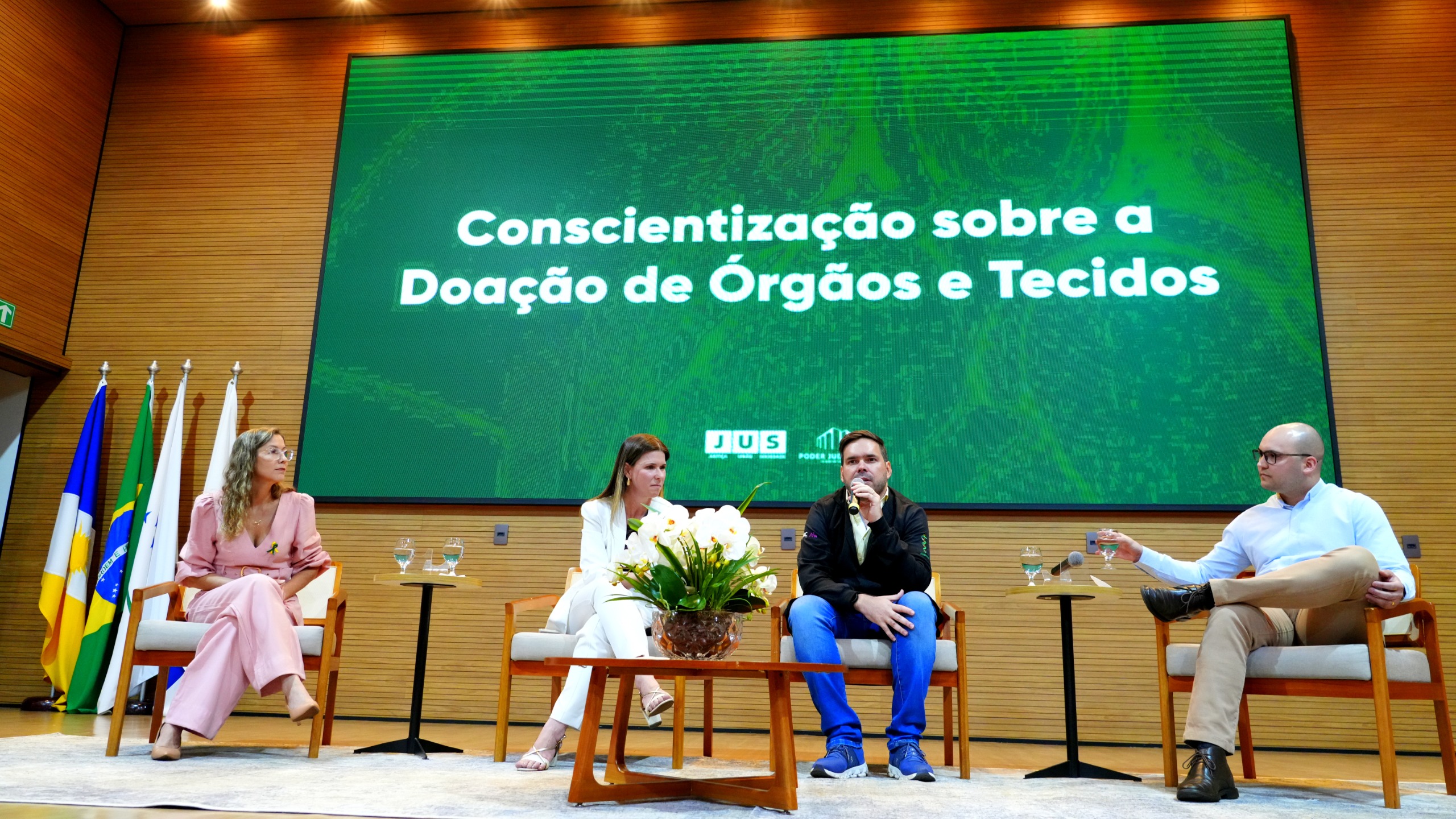 Nas imagens aparcem quatro palestrantes estão sentados em poltronas de madeira clara, dispostas em semicírculo sobre o palco. À esquerda, uma mulher de macacão rosa, ao centro outra mulher de terno branco e, à direita, dois homens: um de jaqueta preta com microfone na mão e outro de camisa branca e calça bege. Entre eles, uma mesa de centro com vaso de orquídeas brancas. Ao fundo, grande tela verde exibe: “Conscientização sobre a Doação de Órgãos e Tecidos”. À esquerda do palco, aparecem as bandeiras do Brasil, do Tocantins e do TJTO. O ambiente é formal e organizado.