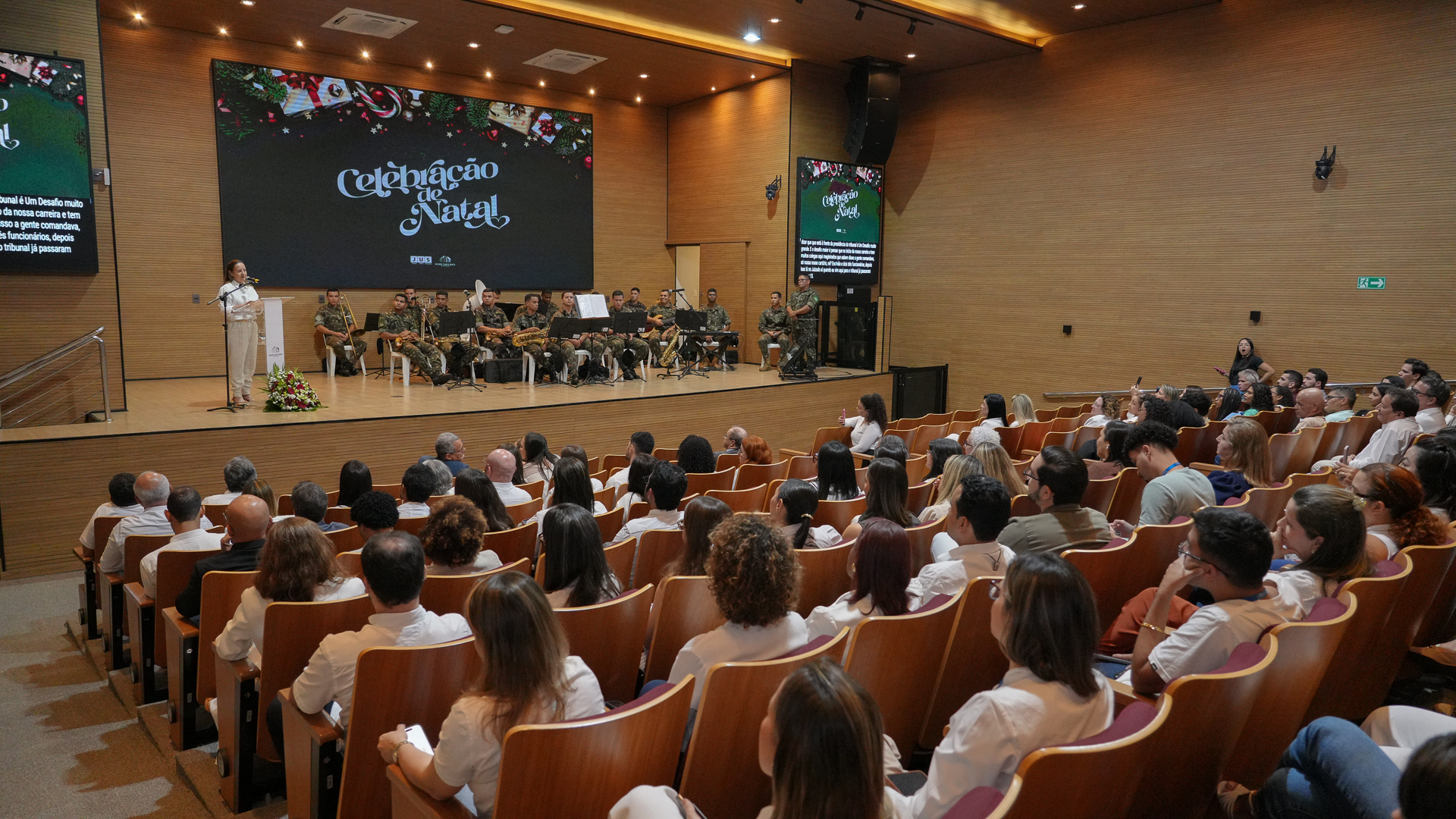 Auditório amplo e moderno, lotado de servidores e servidoras sentados, assistindo à Celebração de Natal do TJTO. No palco, à esquerda, uma mulher fala ao púlpito; ao fundo, uma banda militar, fardada, está posicionada com instrumentos musicais. Telões laterais e o painel central exibem a identidade visual do evento com o título “Celebração de Natal”. O clima é solene e acolhedor, com decoração natalina projetada nos displays e iluminação quente que valoriza o ambiente.