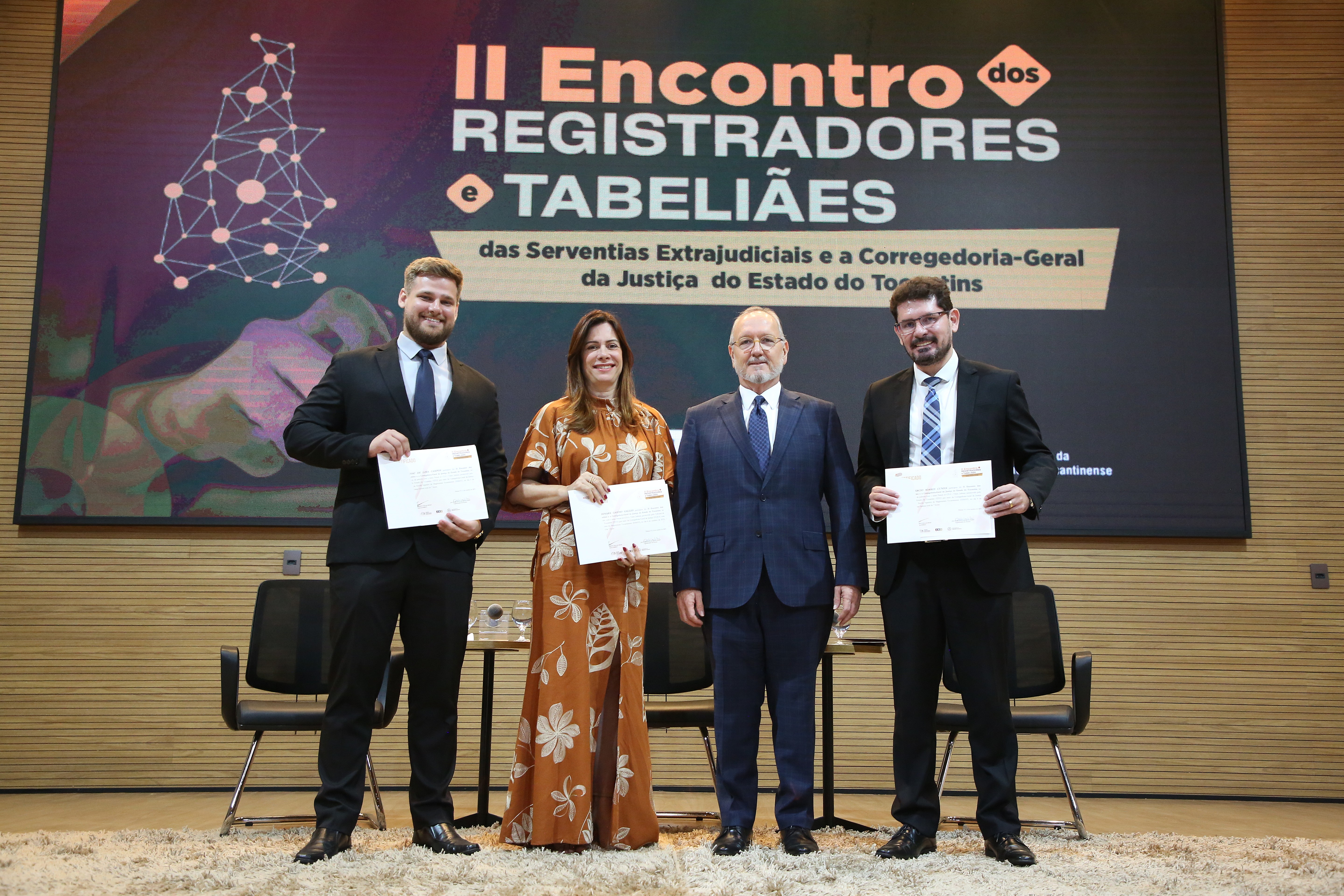 A imagem mostra quatro pessoas sorrindo e segurando diplomas em um evento formal diante de um banner do “II Encontro dos Registradores e Tabeliães”, simbolizando reconhecimento e celebração.