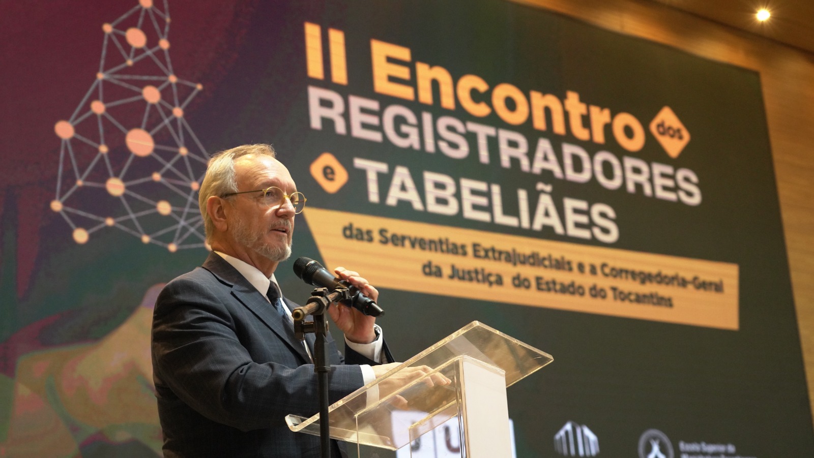 A imagem mostra um homem de terno e óculos discursando com microfone em um púlpito, diante de uma tela que anuncia o “II Encontro dos Registradores e Tabeliães”, em ambiente formal e iluminado.