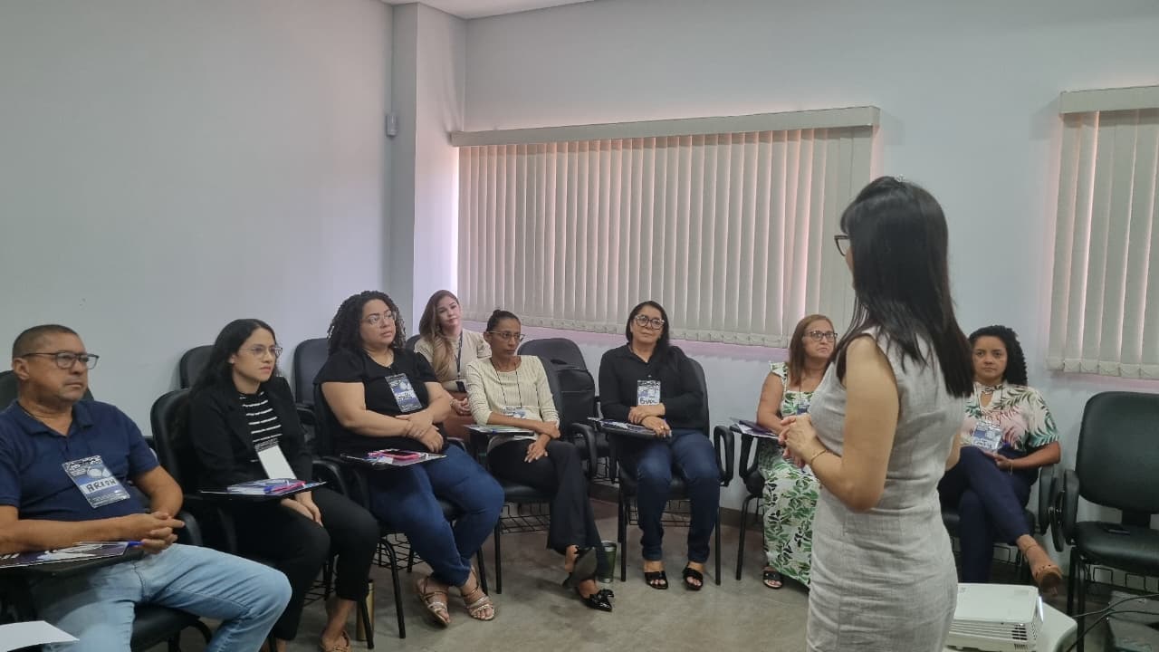 Em uma sala de reuniões de paredes claras, com persianas fechadas, um grupo de oito pessoas está sentado em cadeiras com apoio para anotações, formando um semicírculo. Elas observam atentamente as explicações que estão sendo feitas por uma mulher de cabelos lisos e escuros, de vestido cinza claro sem mangas, que parece conduzir a atividade ou falar ao grupo.