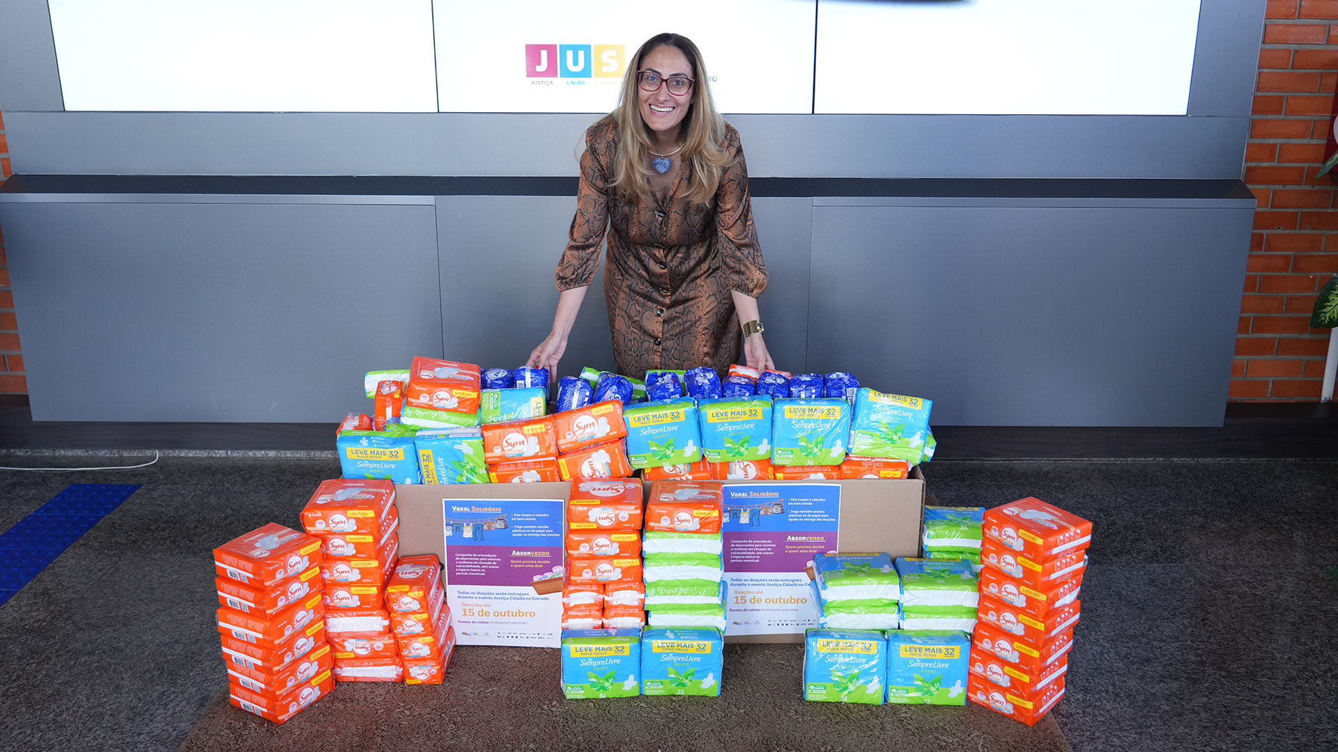Juíza Rosa Maria Gazire Rossi, sorridente, de pé, atrás de vários pacotes de absorventes higiênicos de diferentes marcas e cores. Ela veste um vestido marrom estampado e óculos, transmitindo simpatia e acolhimento. 