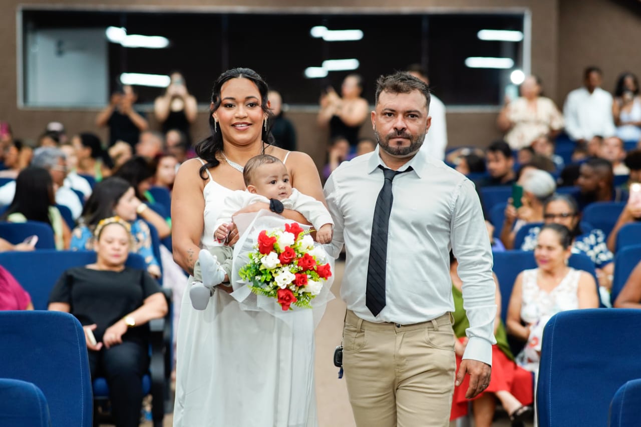 A imagem mostra a família caminhando pelo salão durante o casamento, com os pais sorrindo e segurando o bebê, enquanto convidados sentados em cadeiras azuis acompanham emocionados o momento.