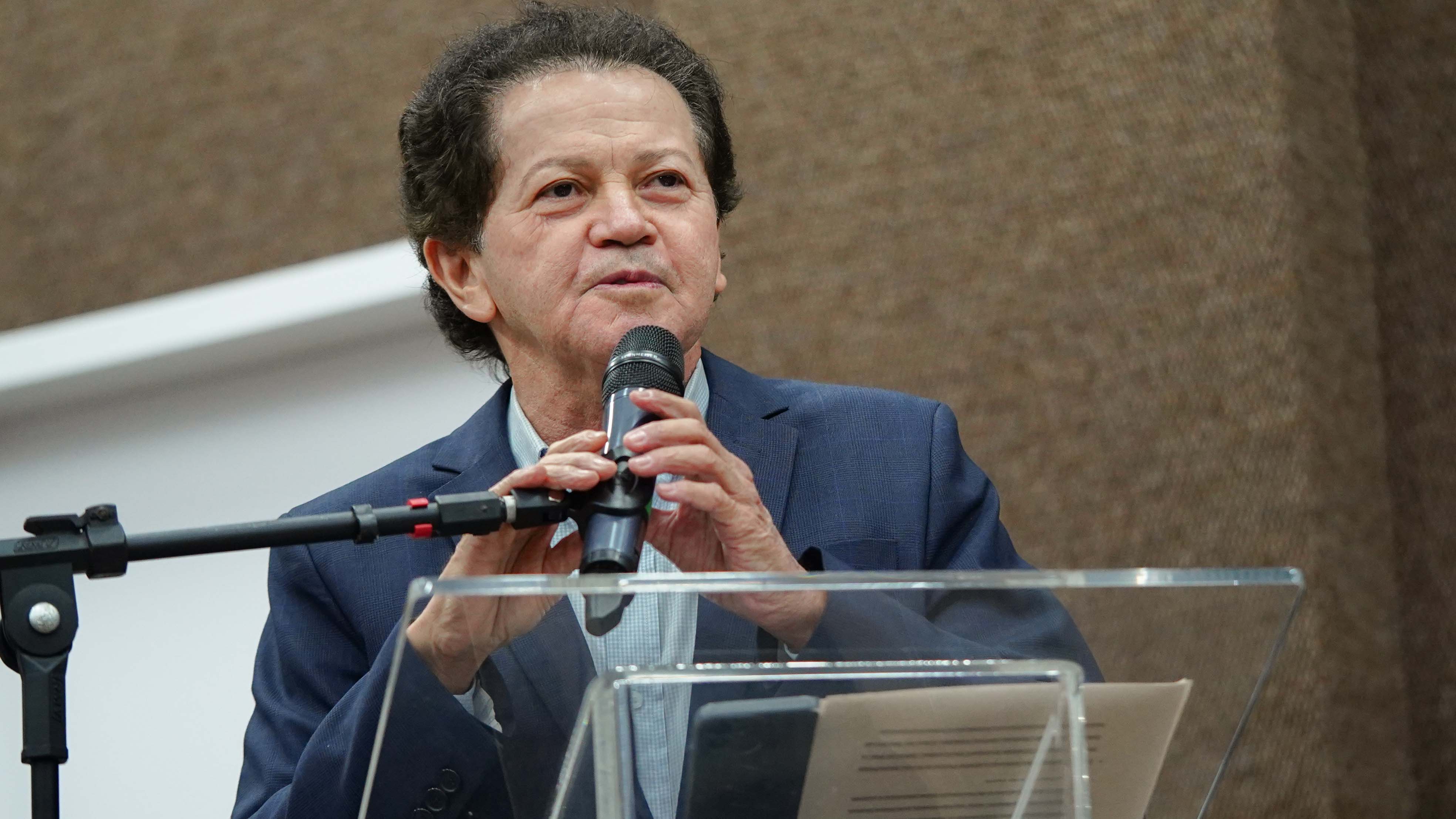 Juiz Deusamar Alves, homem moreno, fala ao microfone, e usa terno e camisa azul