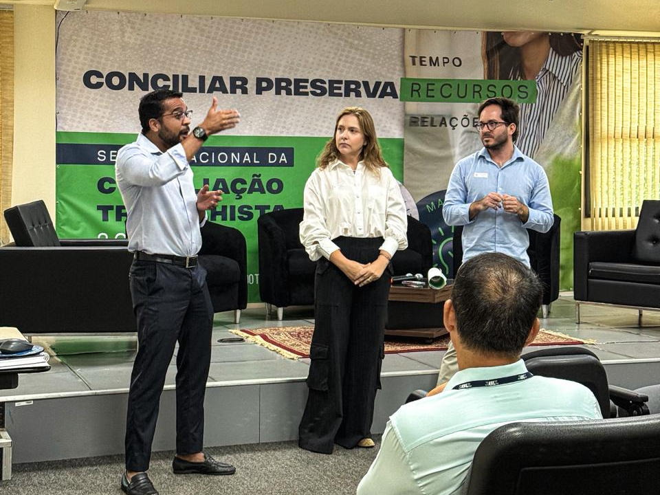 Imagem com três pessoas em pé, conduzindo uma apresentação diante de um público, em um auditório. À esquerda, um homem fala com gestos expressivos; ao centro, uma mulher escuta atentamente, e à direita, outro homem aguarda sua vez de falar, em postura colaborativa. Ao fundo, um painel institucional.