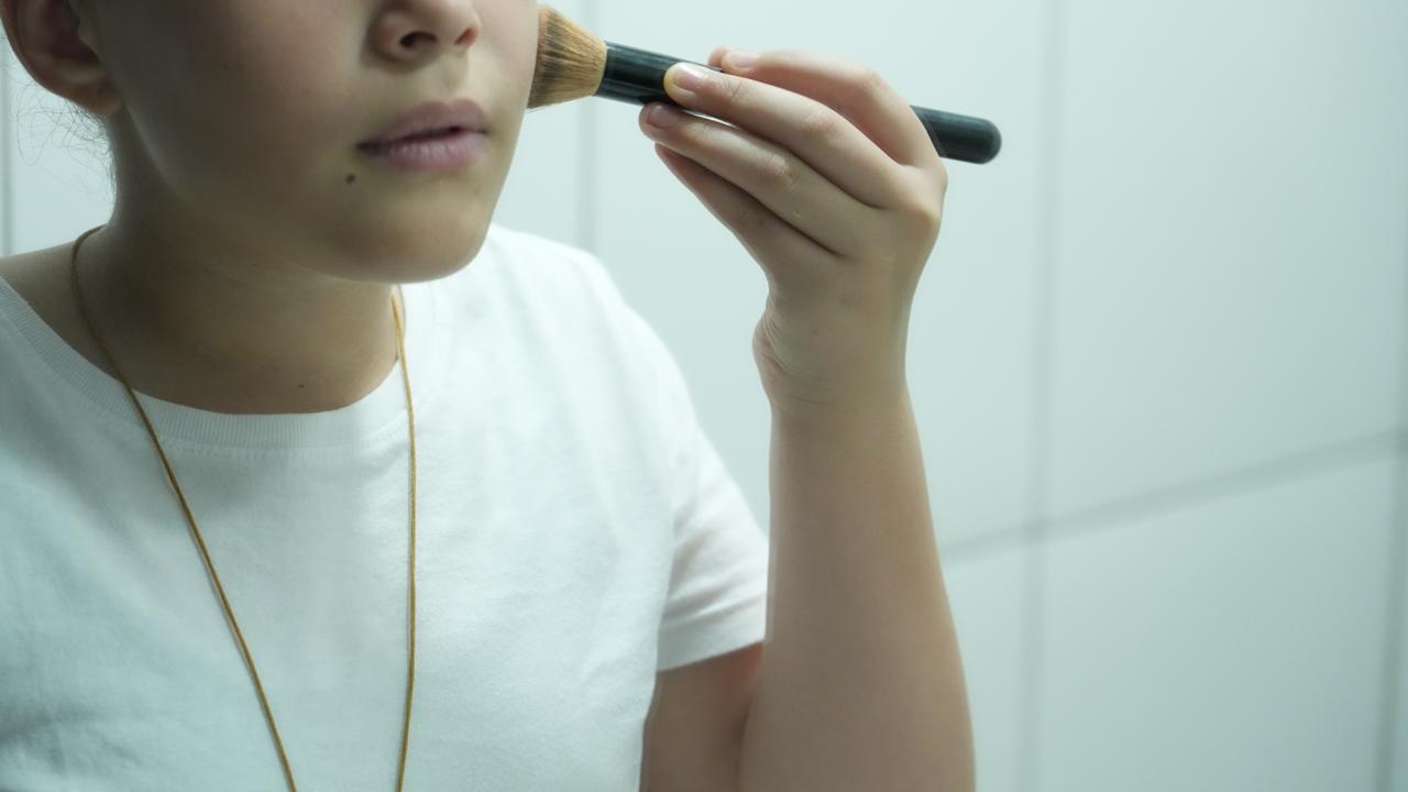 Imagem mostra uma criança aplicando maquiagem com um pincel no rosto, em frente a um espelho